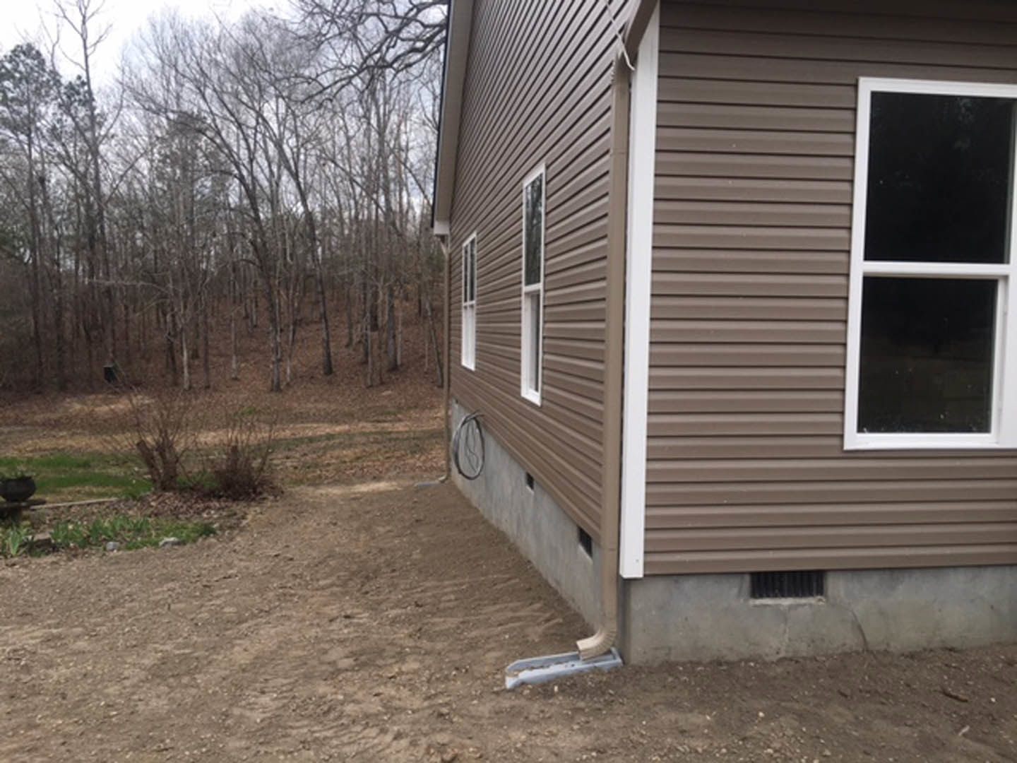 Two-story home with light-colored siding, white-framed windows, and a dirt path leading to the entrance, surrounded by mature trees and greenery in the background.