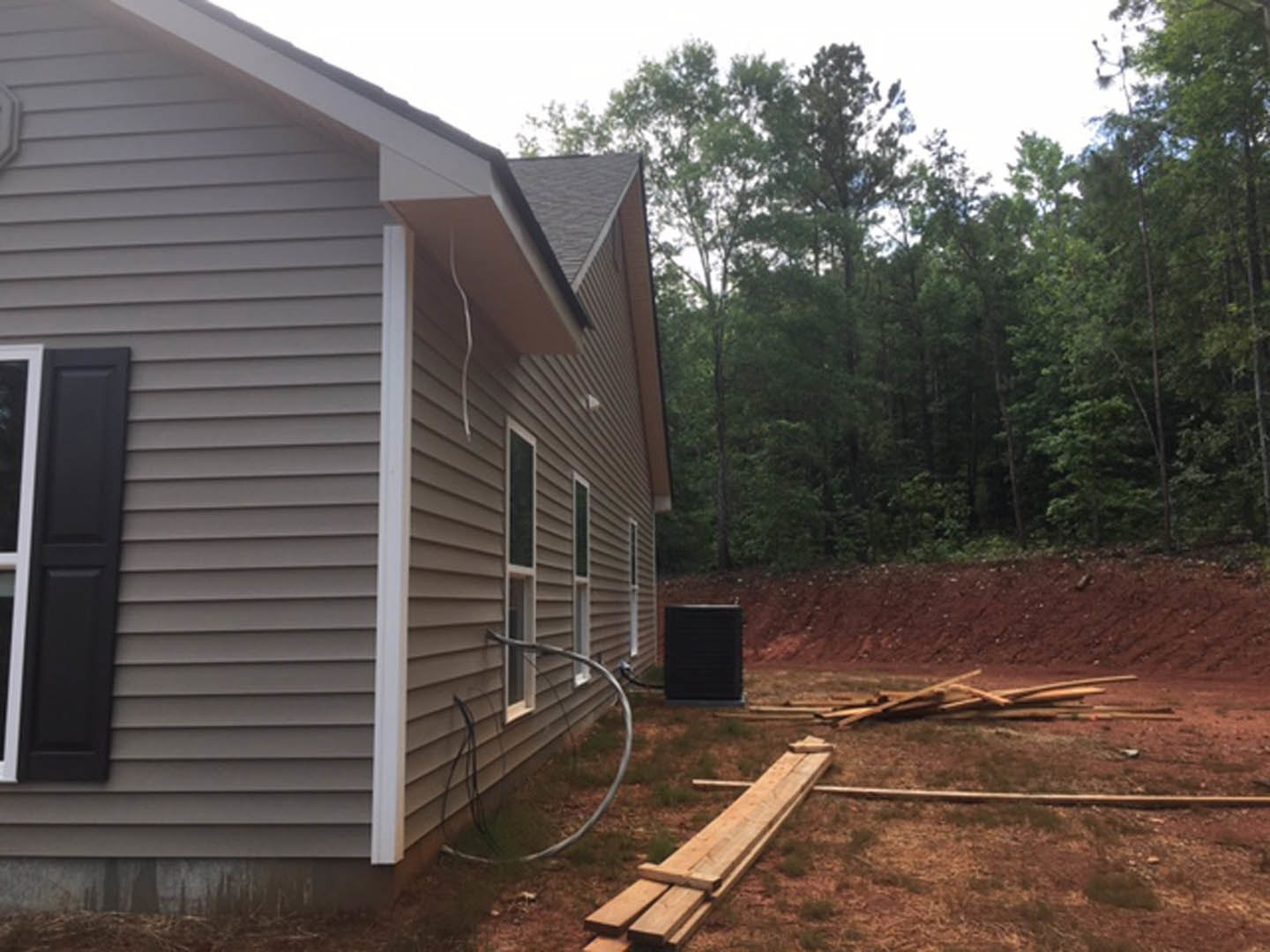 Two-story home with horizontal wood plank siding, black shutters, metal pipe, and grey fence; surrounded by trees and plants under a clear sky.