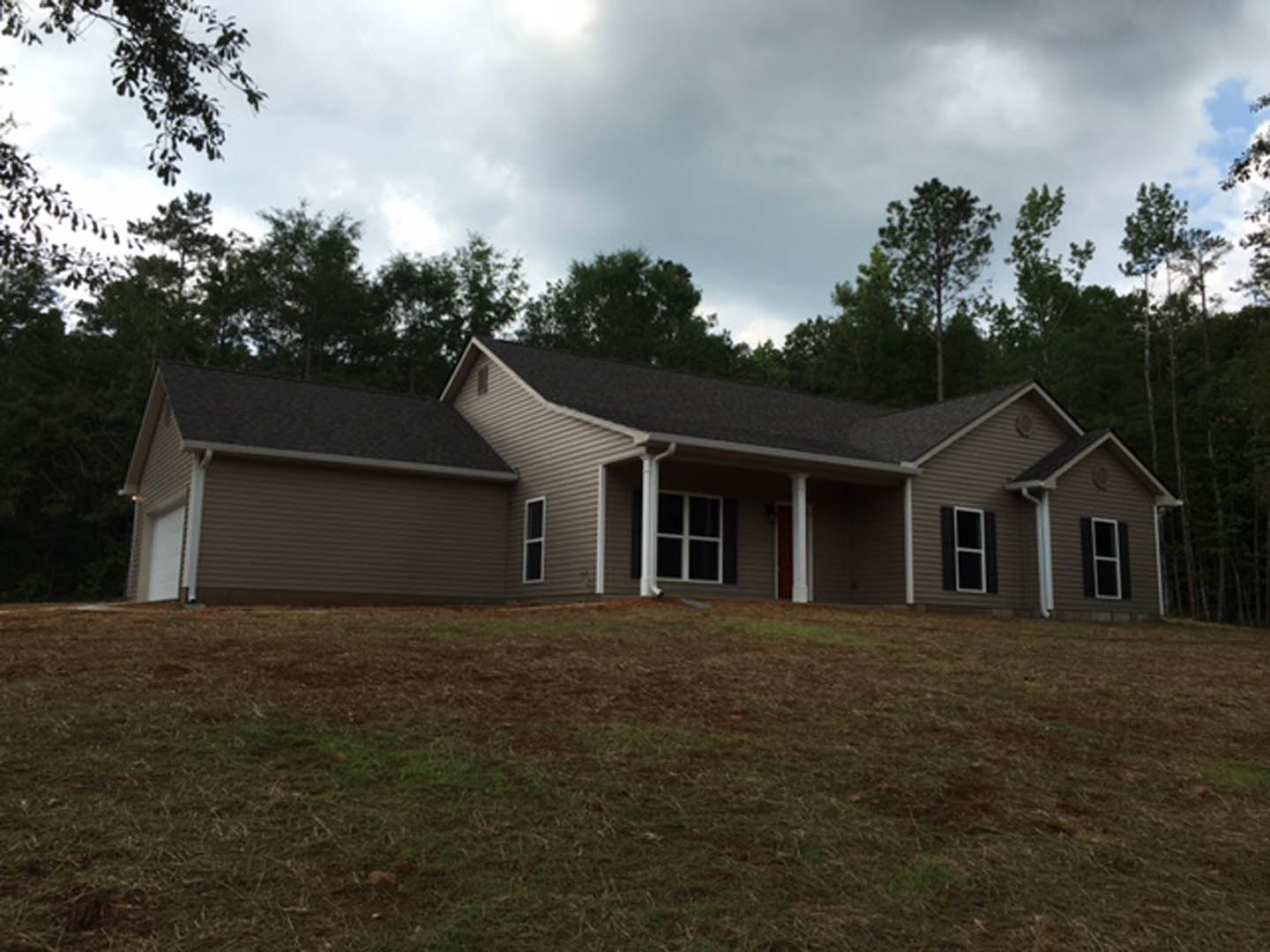 Two-story house with white door and white-framed windows, gray shingle roof, surrounded by large green lawn and mature trees under partly cloudy sky