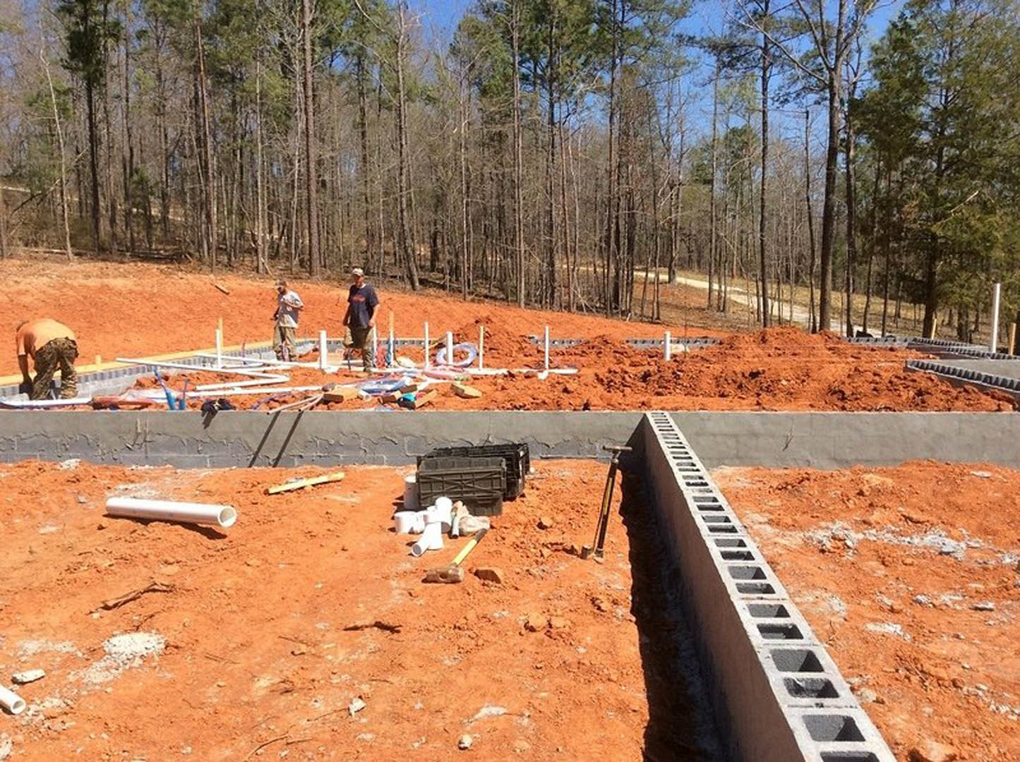 Partially built custom home with exposed concrete block foundation, soil and construction materials in foreground, several people standing near the structure, trees and sky in