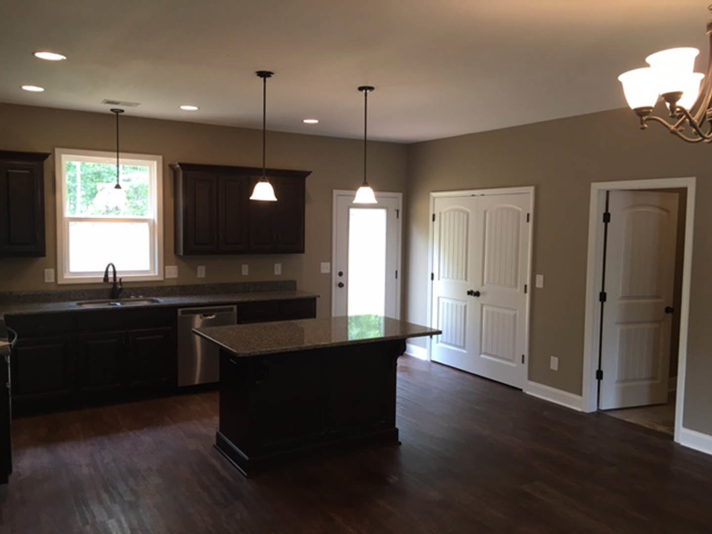 Black kitchen island with white marble countertop, wood flooring, white cabinetry with black hardware, ceiling pendant light, window above sink and faucet, white door with black