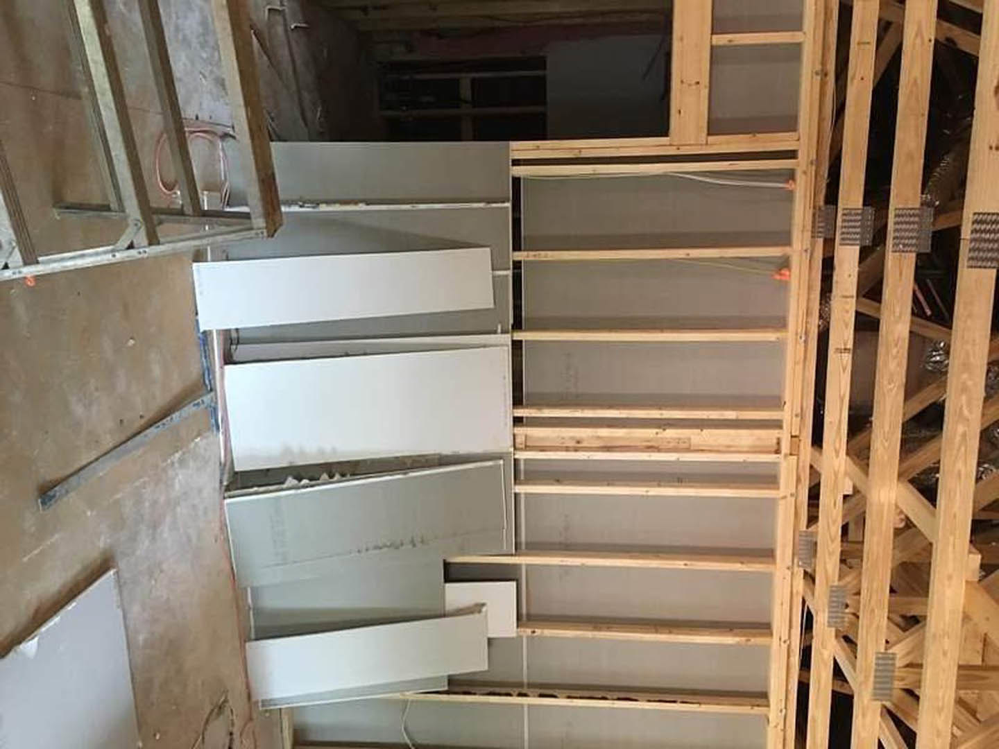 Unfinished room with exposed wood ceiling beams, partially framed walls with white panels, built-in wood shelving, and construction materials scattered throughout.