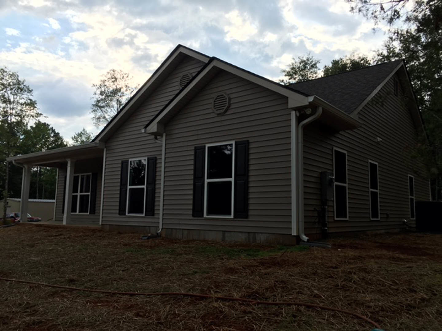 Two-story house with gray siding, white-trimmed windows, gabled roof, grassy yard, garden hose on ground, mature trees in background, cloudy sky overhead.