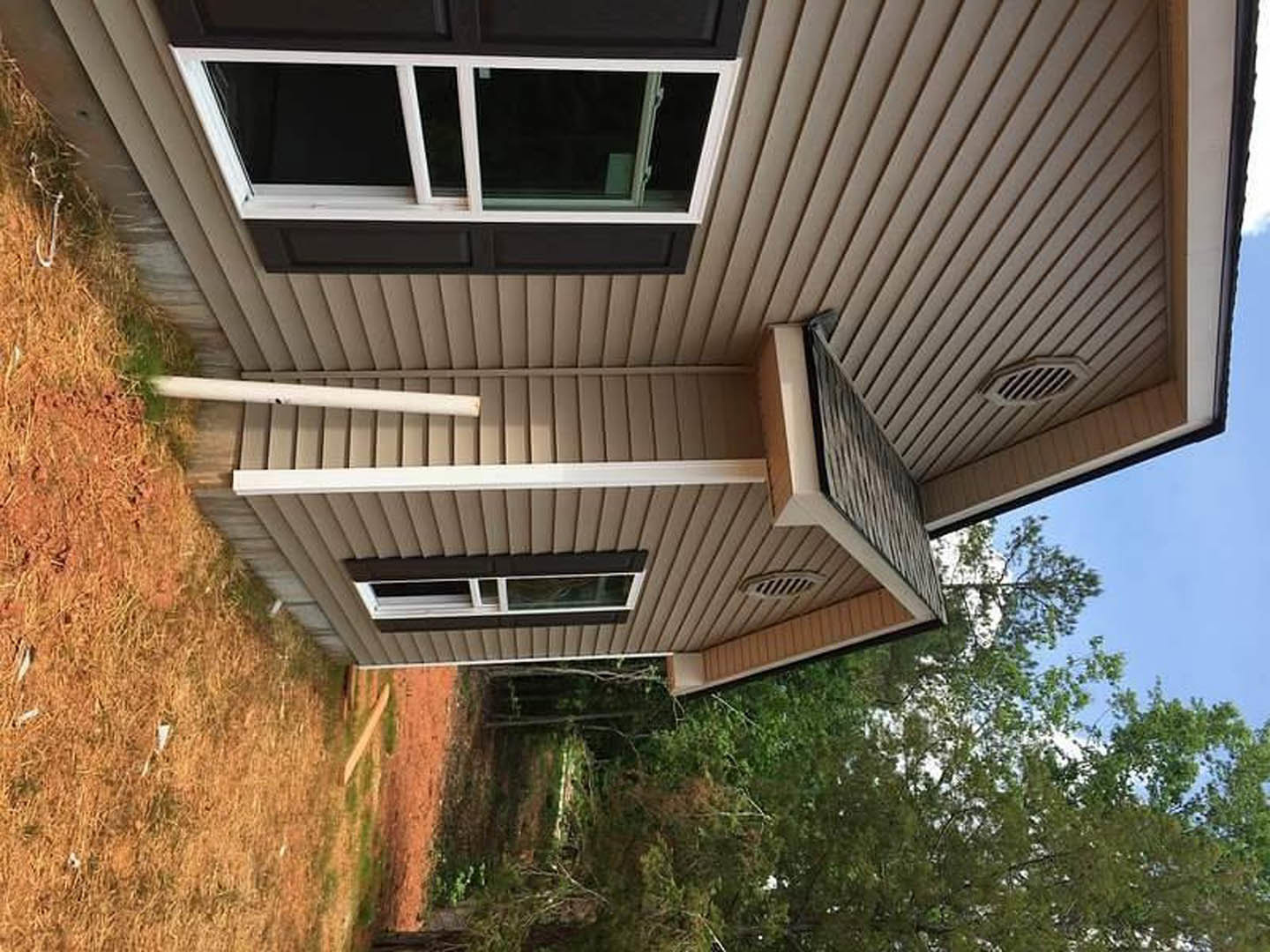 Two-story house with covered porch, white columns, brick chimney, large windows, and balcony railing, surrounded by trees and landscaping.