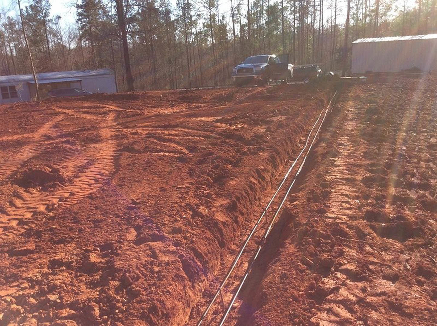 White pickup truck parked on a dirt lot beside young trees, with exposed soil and tire tracks visible under a clear sky.