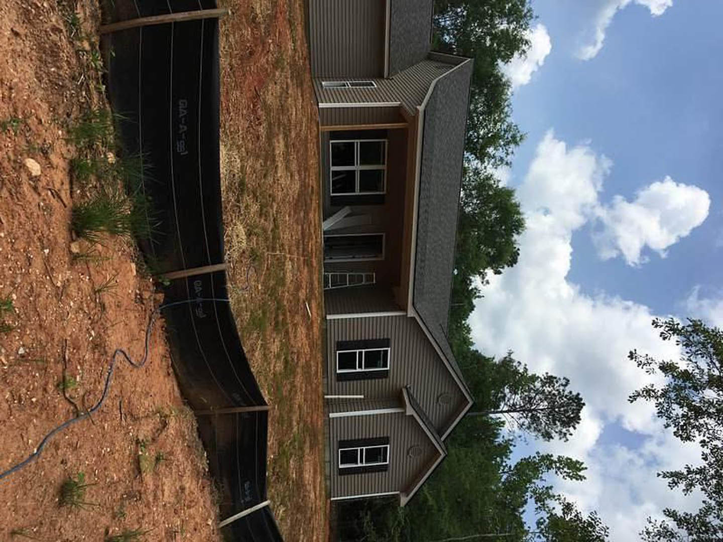 White rectangular window with metal frame set in a modern house exterior, black pipe visible in grassy backyard, wooden fence and trees in the background, partly cloudy sky