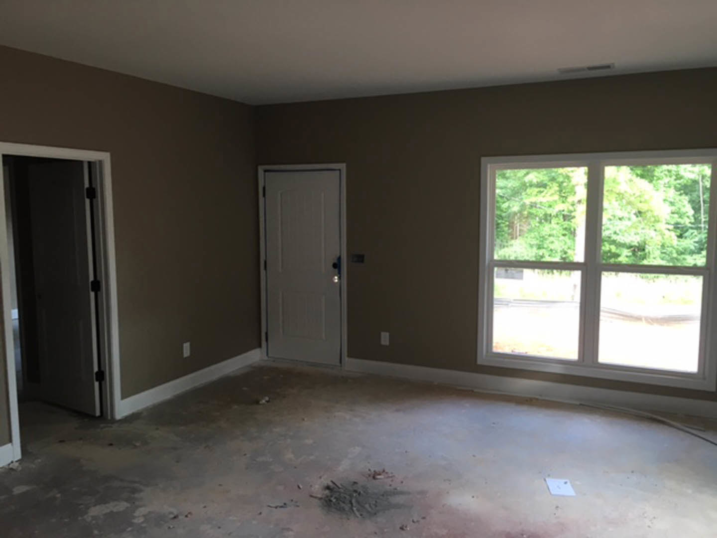 White paneled door with silver knob, rectangular window showing green trees outside, plaster walls, dirty floor scattered with debris