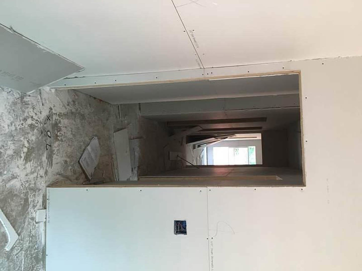 White plaster wall with rectangular hole, light streaming through window, blurred figure partially visible, smooth ceiling, minimal fixtures.