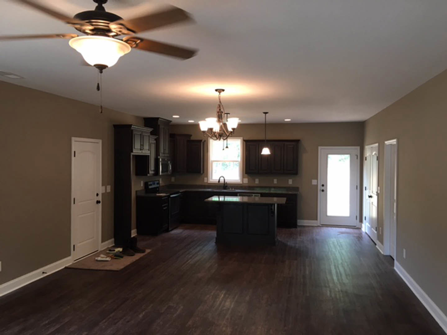 Open kitchen with two ceiling fans, white cabinetry, light countertops, white door with black knobs, wood flooring, and a window with a hanging light fixture.