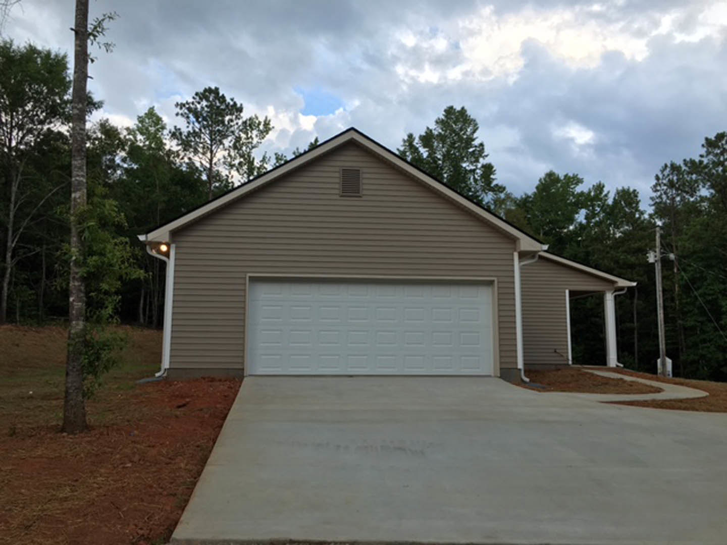 Two-story home with gray siding, attached garage featuring white paneled door, concrete driveway, white columns, and landscaped front yard with trees under a partly cloudy sky