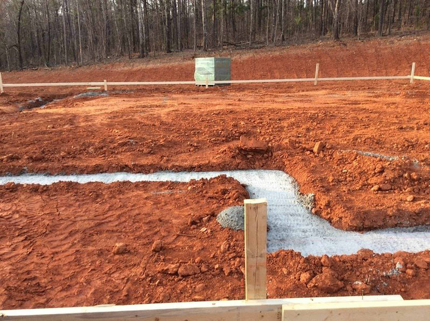 Construction site with wooden fence, exposed soil, tree line in background, and concrete footings visible on ground