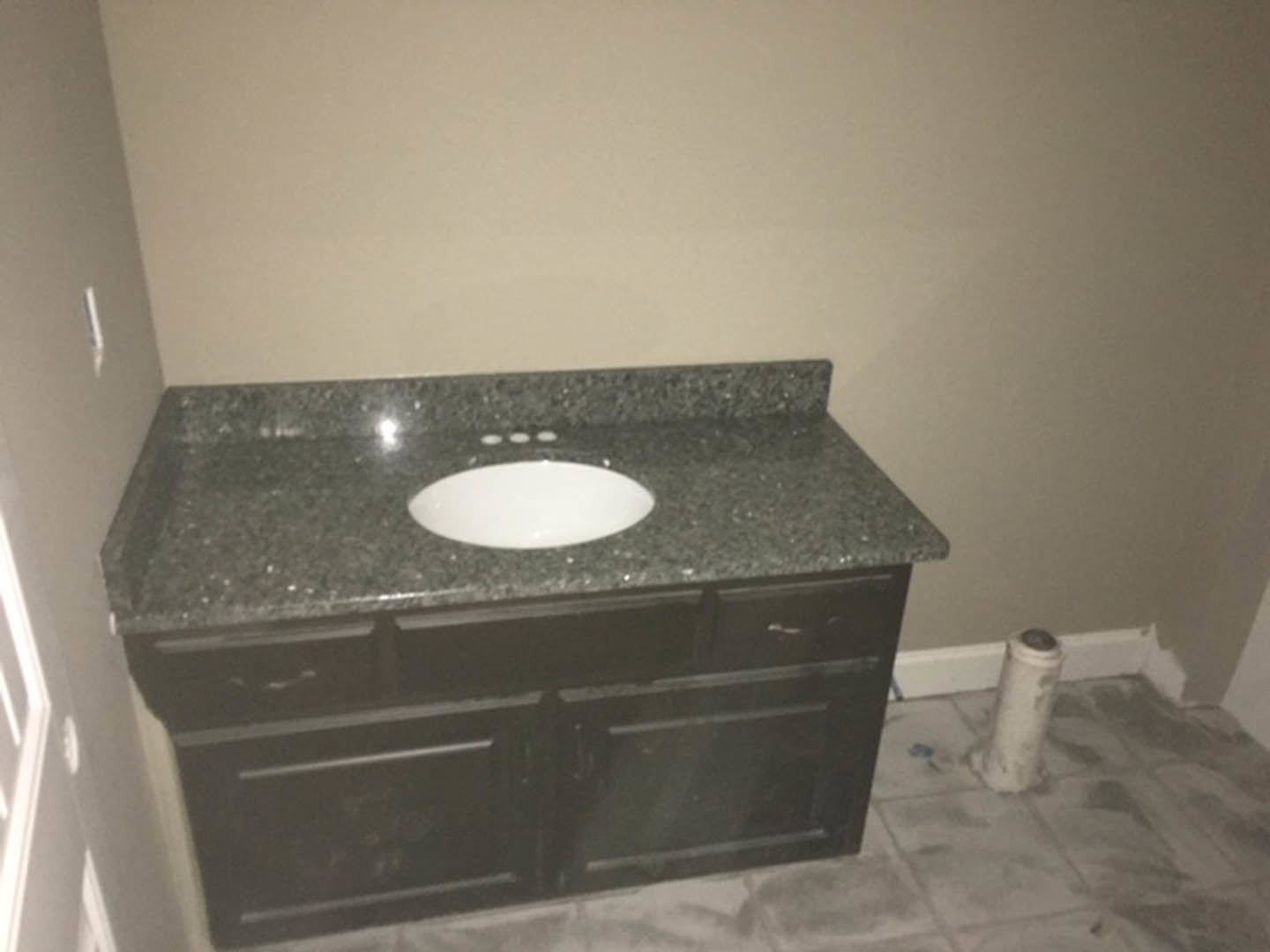 White ceramic vessel sink with chrome faucet set on black stone countertop, black vanity cabinet below, light gray tile backsplash, and white walls in modern bathroom.