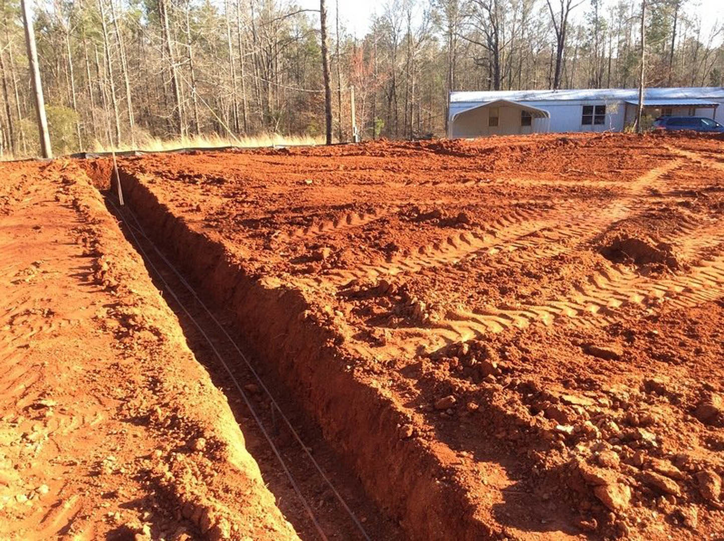 Dirt field with tire tracks and a trench, white trailer parked near trees, partial view of house window with multiple panes