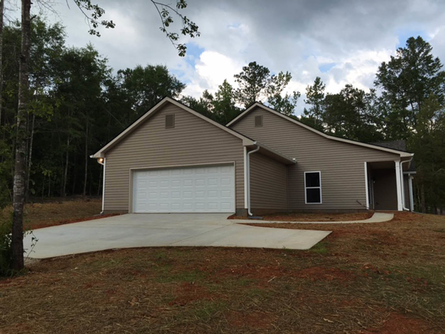 White garage door with brown trim, concrete driveway bordered by grass lawn, white-framed window, mature trees surrounding exterior, light siding and brown accents