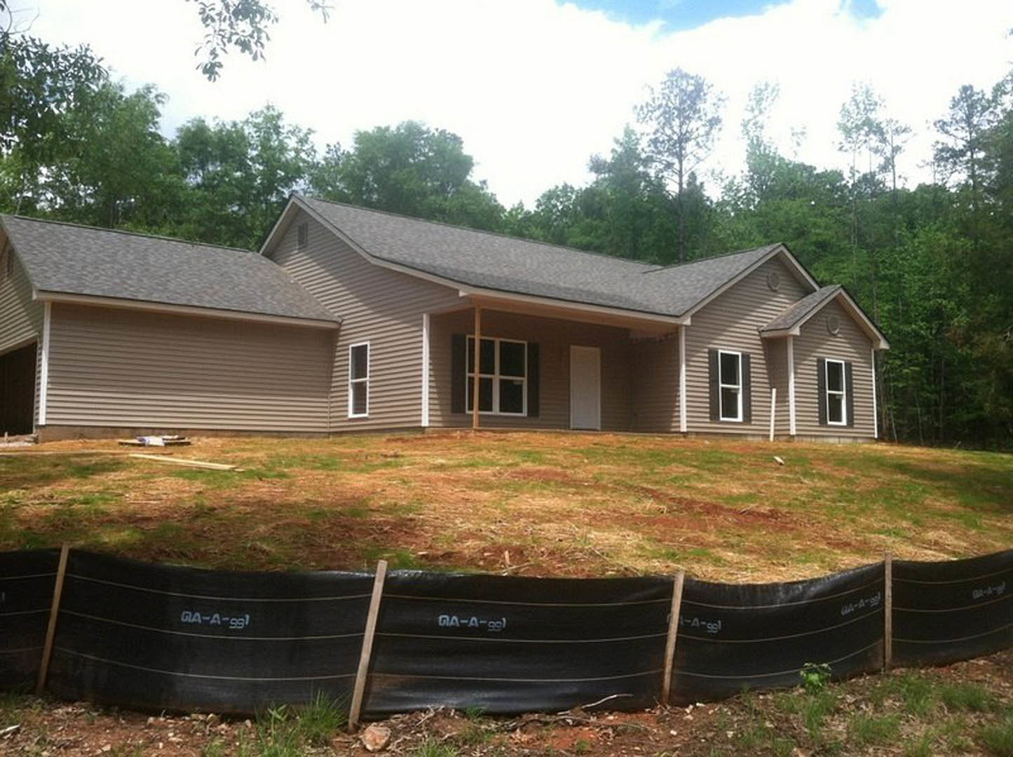 Partially built house with exposed framing and white siding, surrounded by tall trees and grass, black rubber tubing in foreground, small white cottage visible in background.