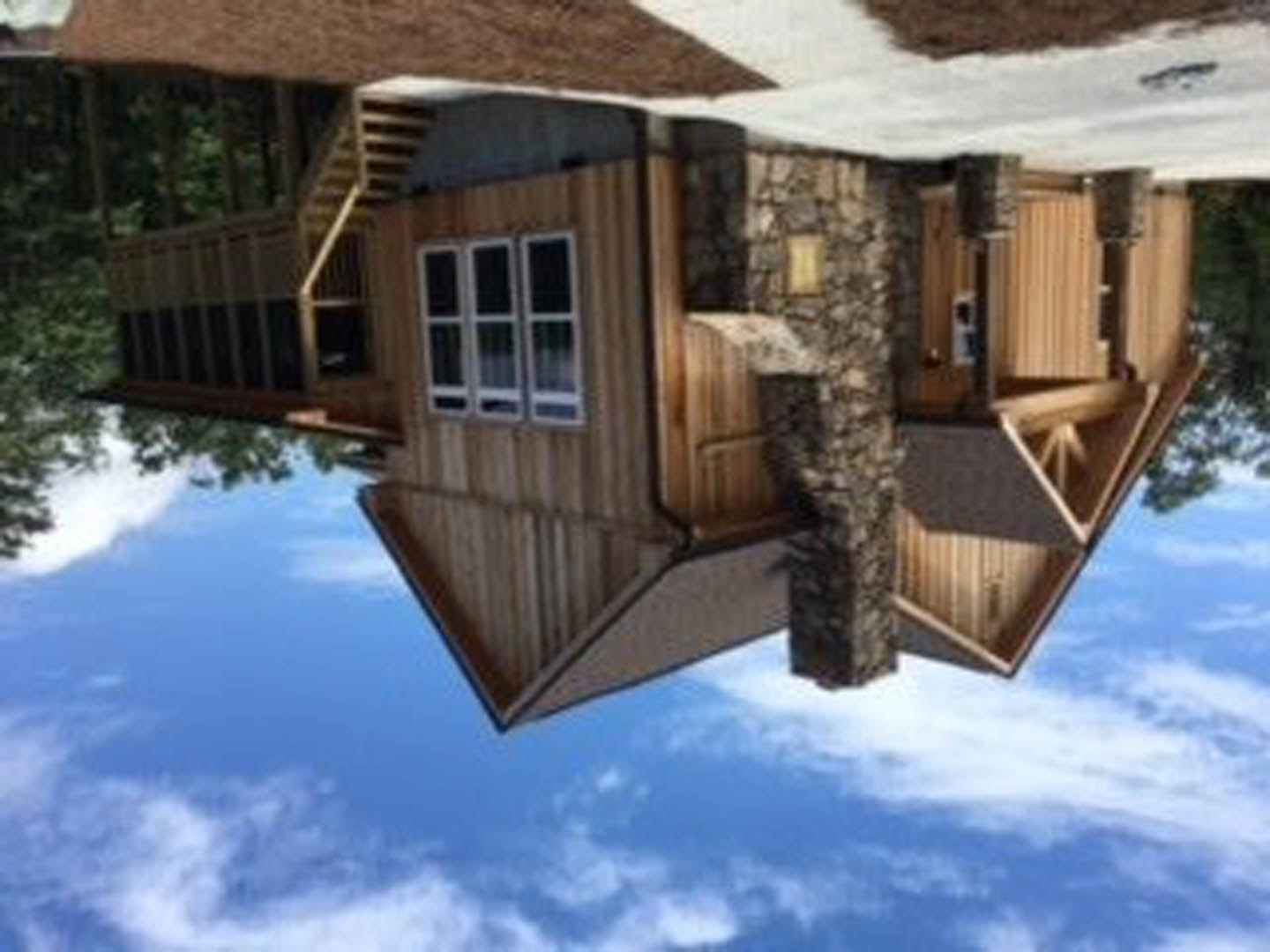 Two-story house with stone walls, large windows, and a wooden staircase, surrounded by trees under a cloudy sky