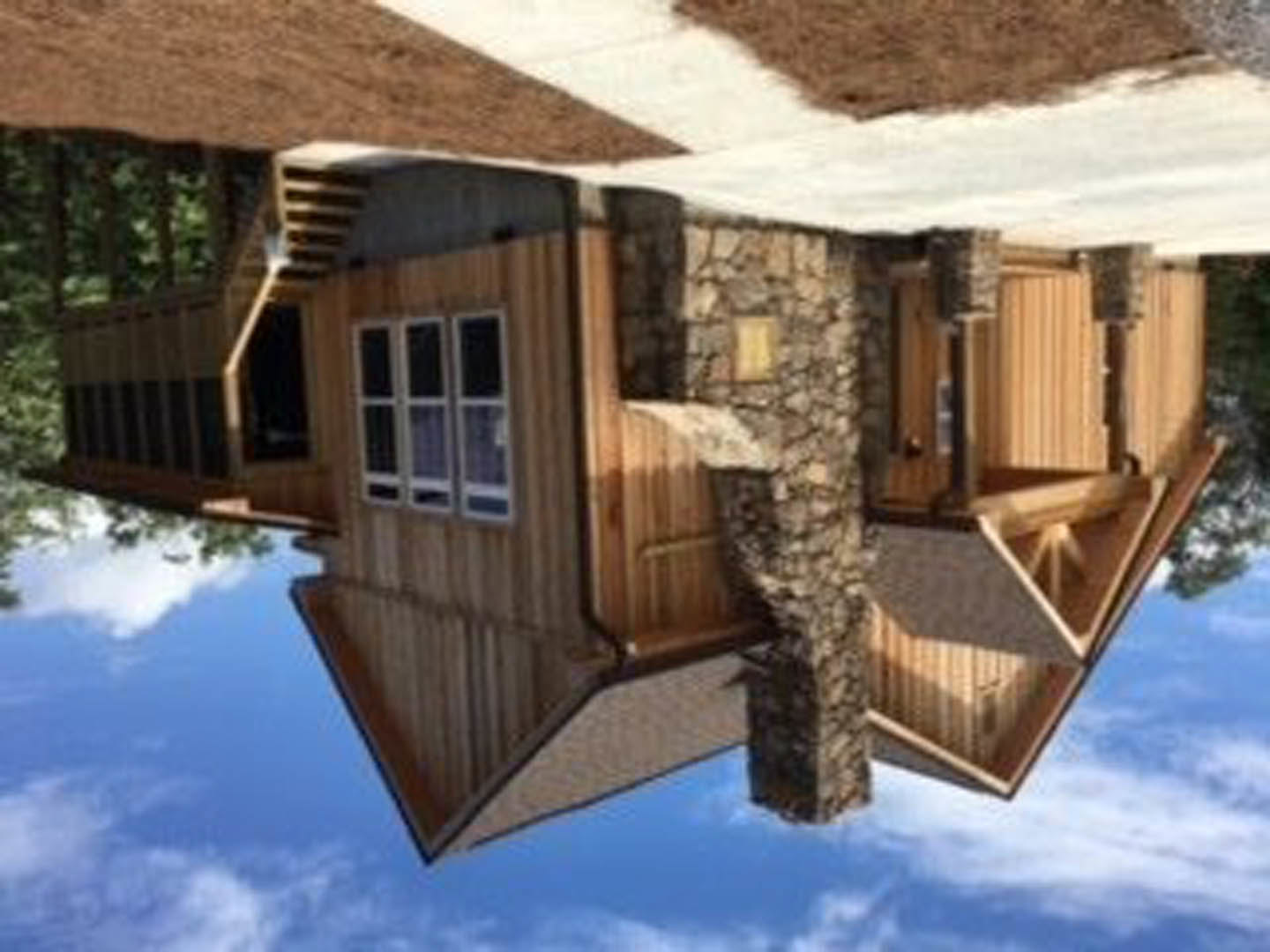 Two-story log cabin with stone chimney, white-trimmed windows, brown wood siding, and mature tree in backyard under cloudy sky
