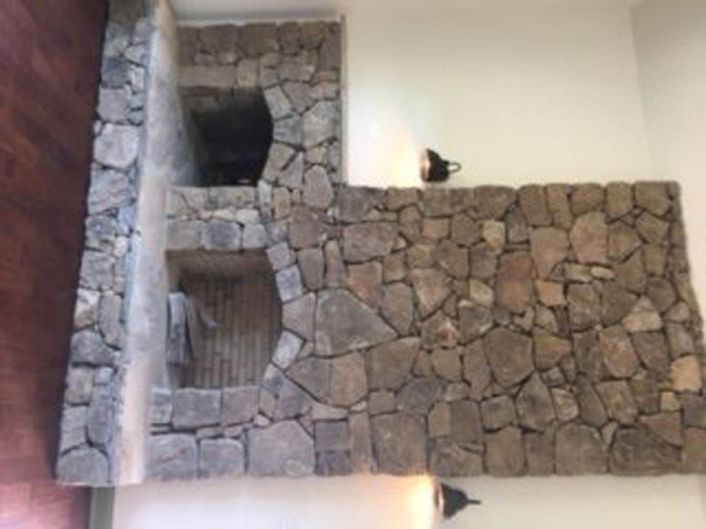 Stone fireplace set into a textured stone wall, surrounded by hardwood floors and neutral painted walls in a spacious living room