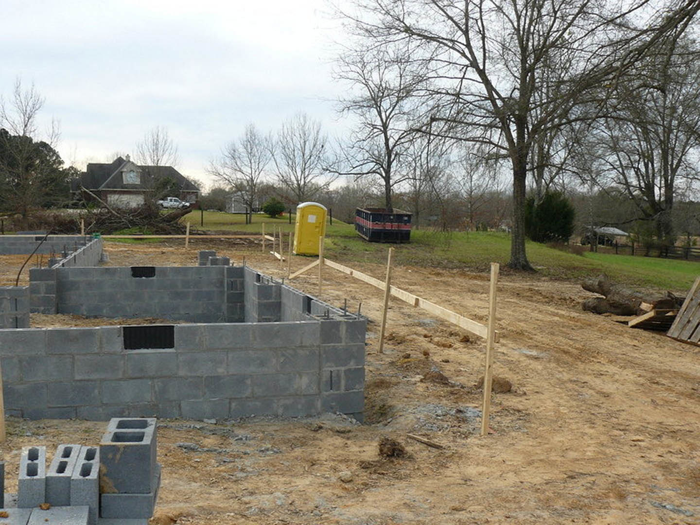 Concrete foundation on grassy building site with yellow portable toilet, scattered construction materials, and trees in background