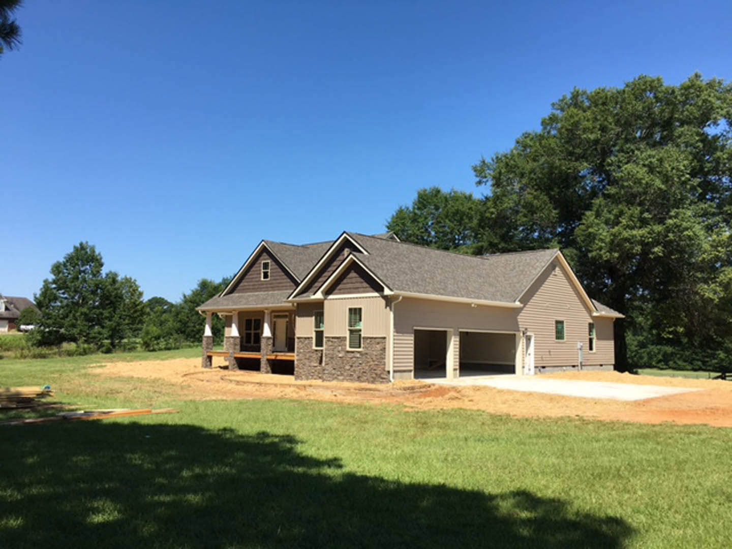 Framed house under construction with exposed wood, attached garage, dirt lot, and mature trees in the background
