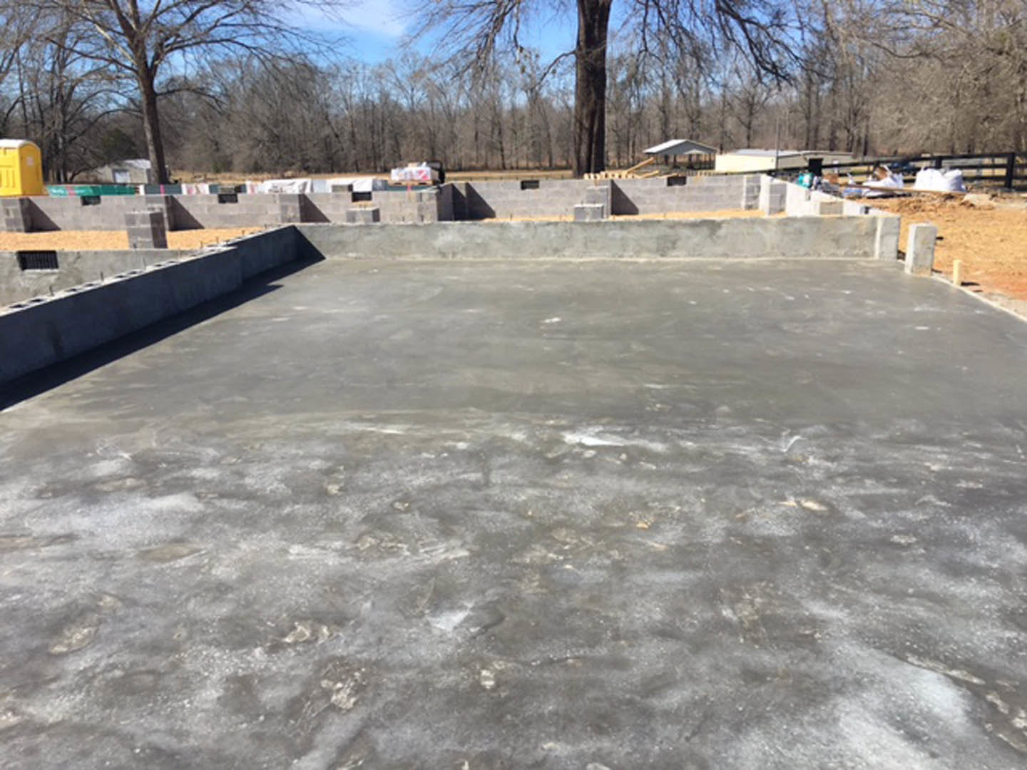 Concrete foundation with stacked concrete blocks, yellow portable toilet on street, trees and sky in background, unfinished construction site
