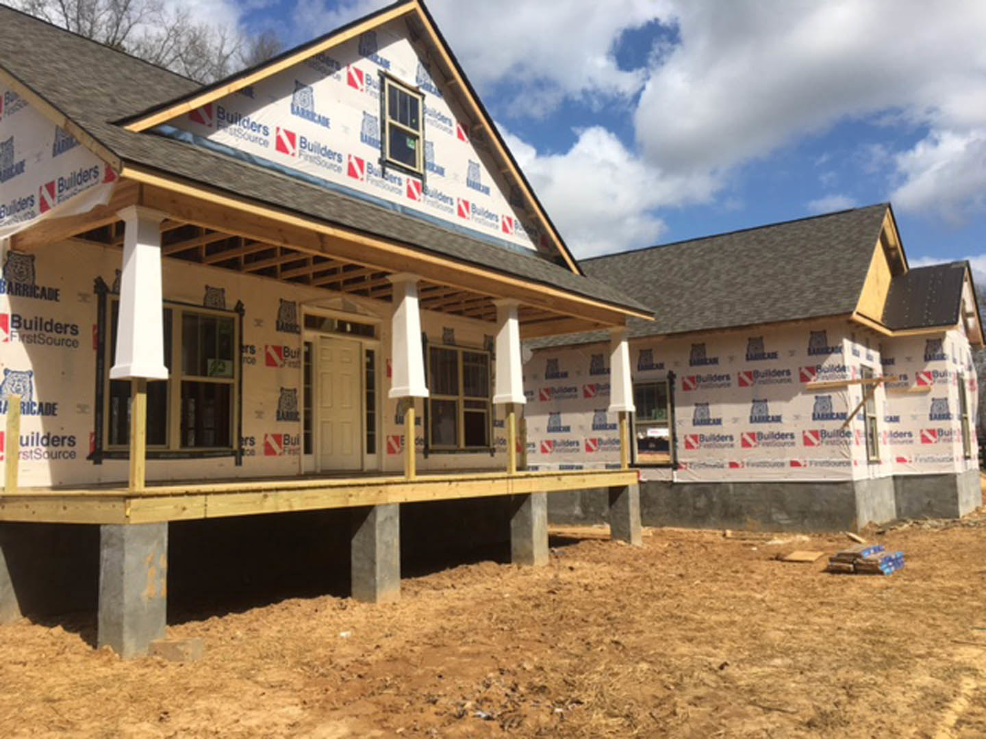 Partially built house with exposed wooden porch, white door with window, white-framed windows, wooden platform foundation, neighboring houses, blue sky with scattered clouds