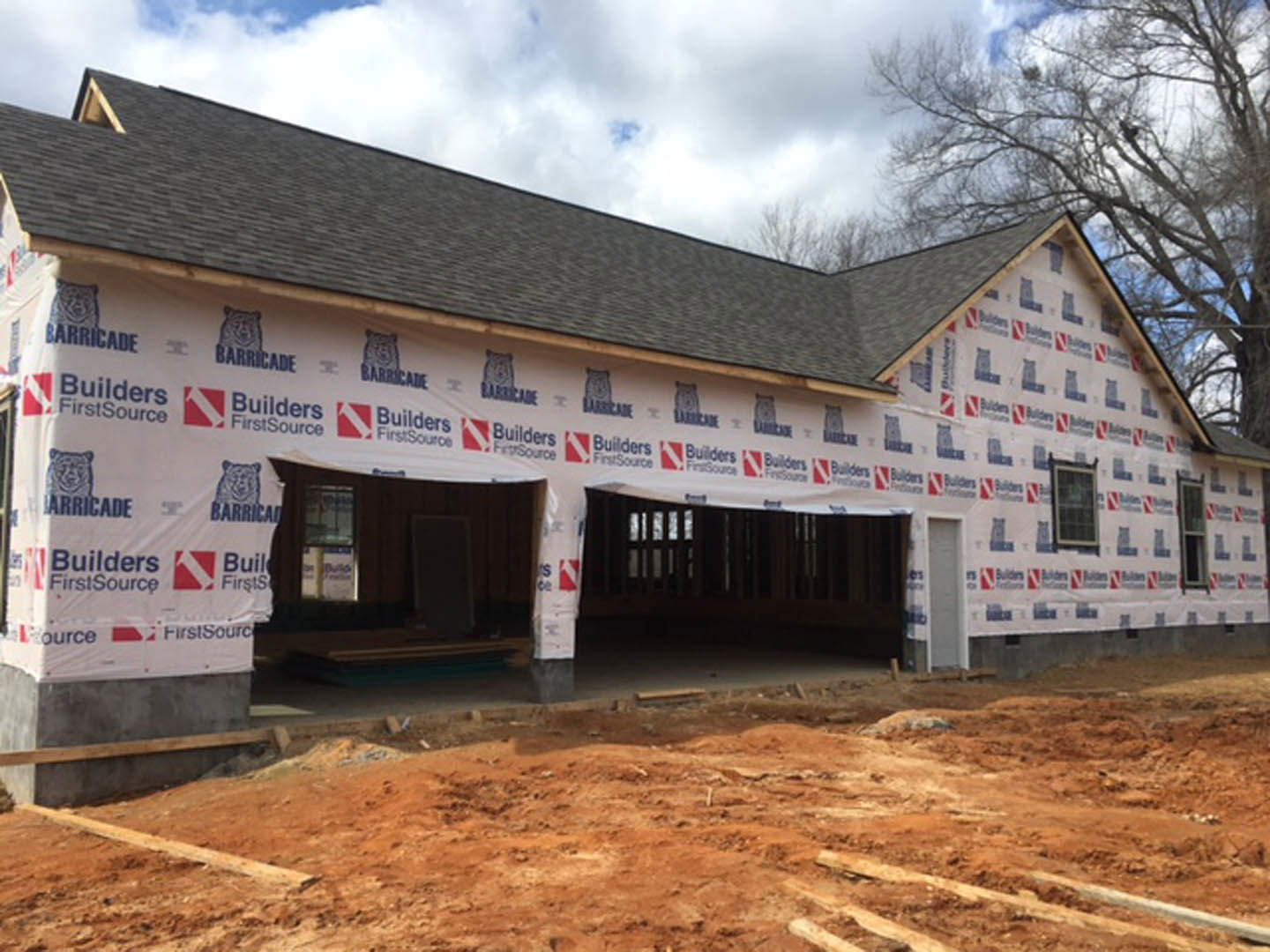 Framed house with attached garage, roof covered in protective sheeting, dirt ground in foreground, partially installed windows, surrounded by trees under cloudy sky