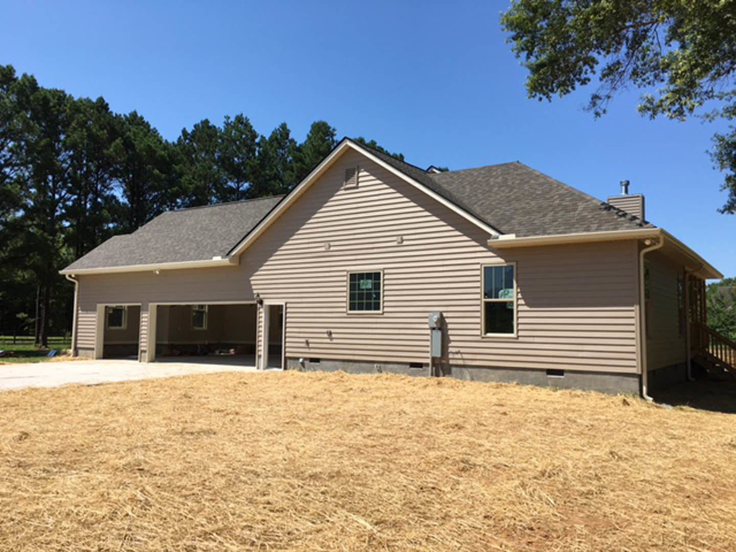 Two-story house with white siding, attached garage, several windows, and a large grassy lawn in front, bordered by mature trees under a clear sky