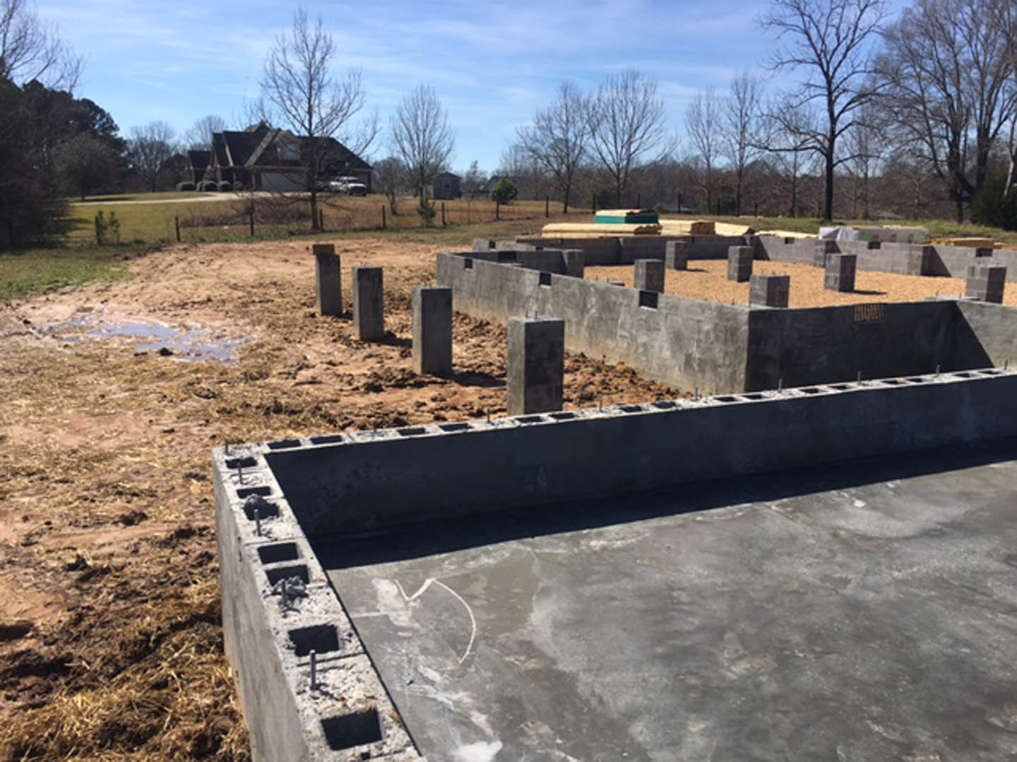 Concrete foundation with vertical pillars and exposed dirt in front of a house, leafless tree and group of trees in the background