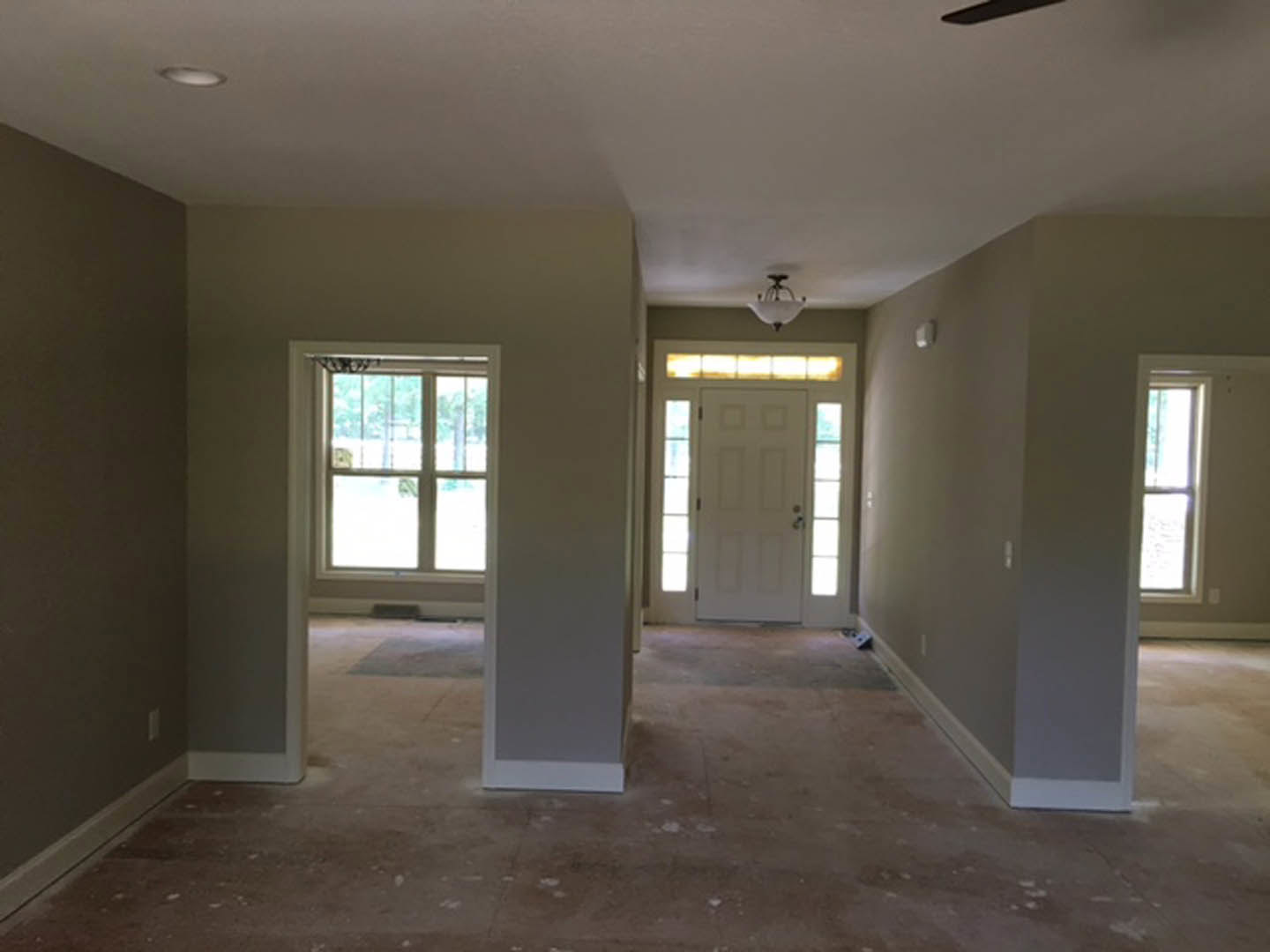White paneled door with silver handle, light fixture on smooth white ceiling, large window letting sunlight onto hardwood floor, neutral plaster walls