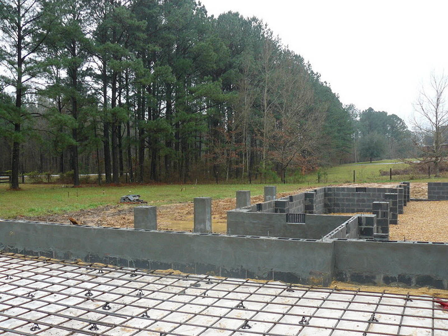 Concrete foundation with exposed metal grid at a residential building site, surrounded by leafless trees and dense forest in the background, grey utility box in grassy area
