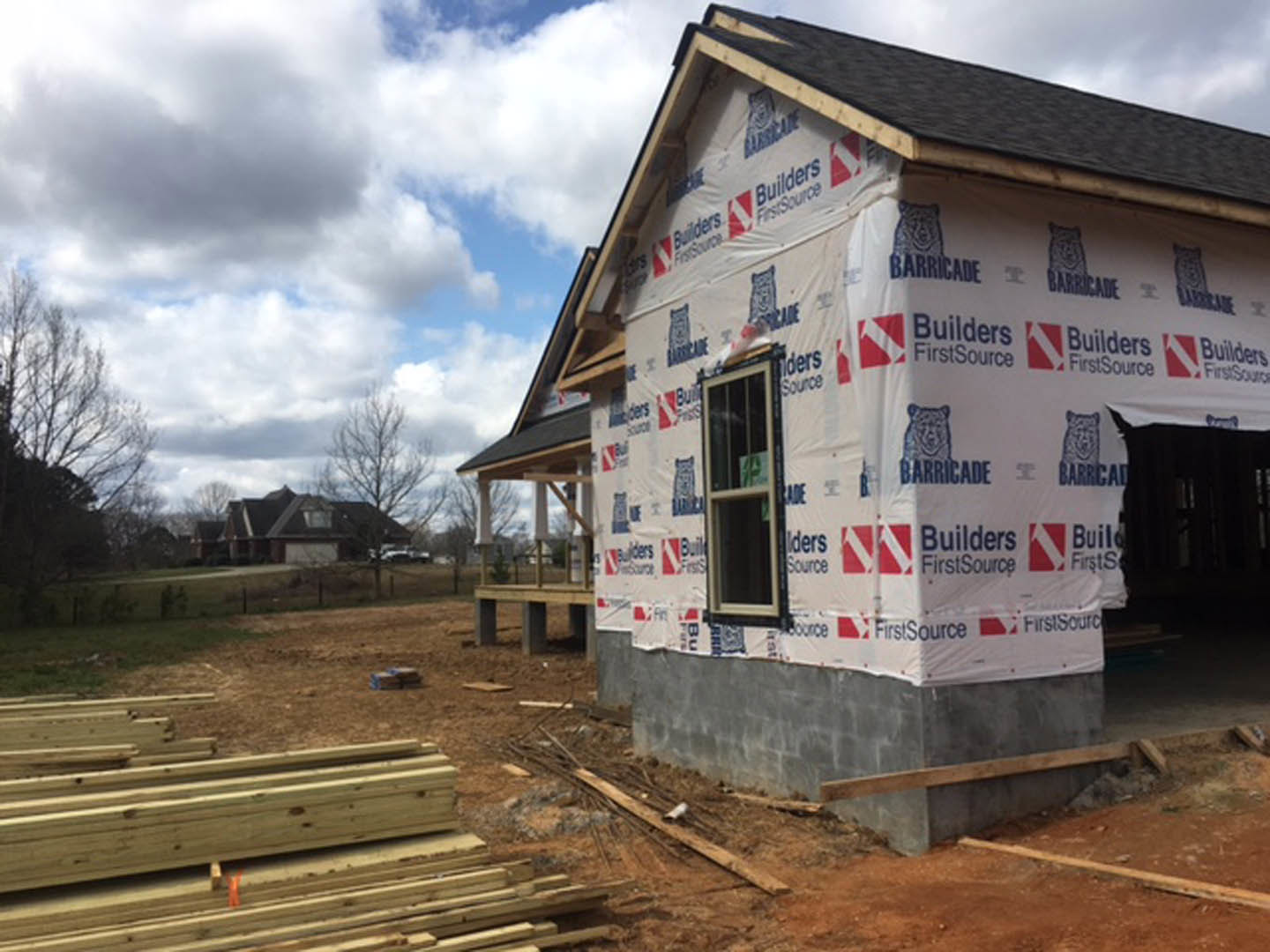 Partially built house with exposed wooden framing, covered porch, pile of lumber on dirt ground, cloudy sky, and trees in background