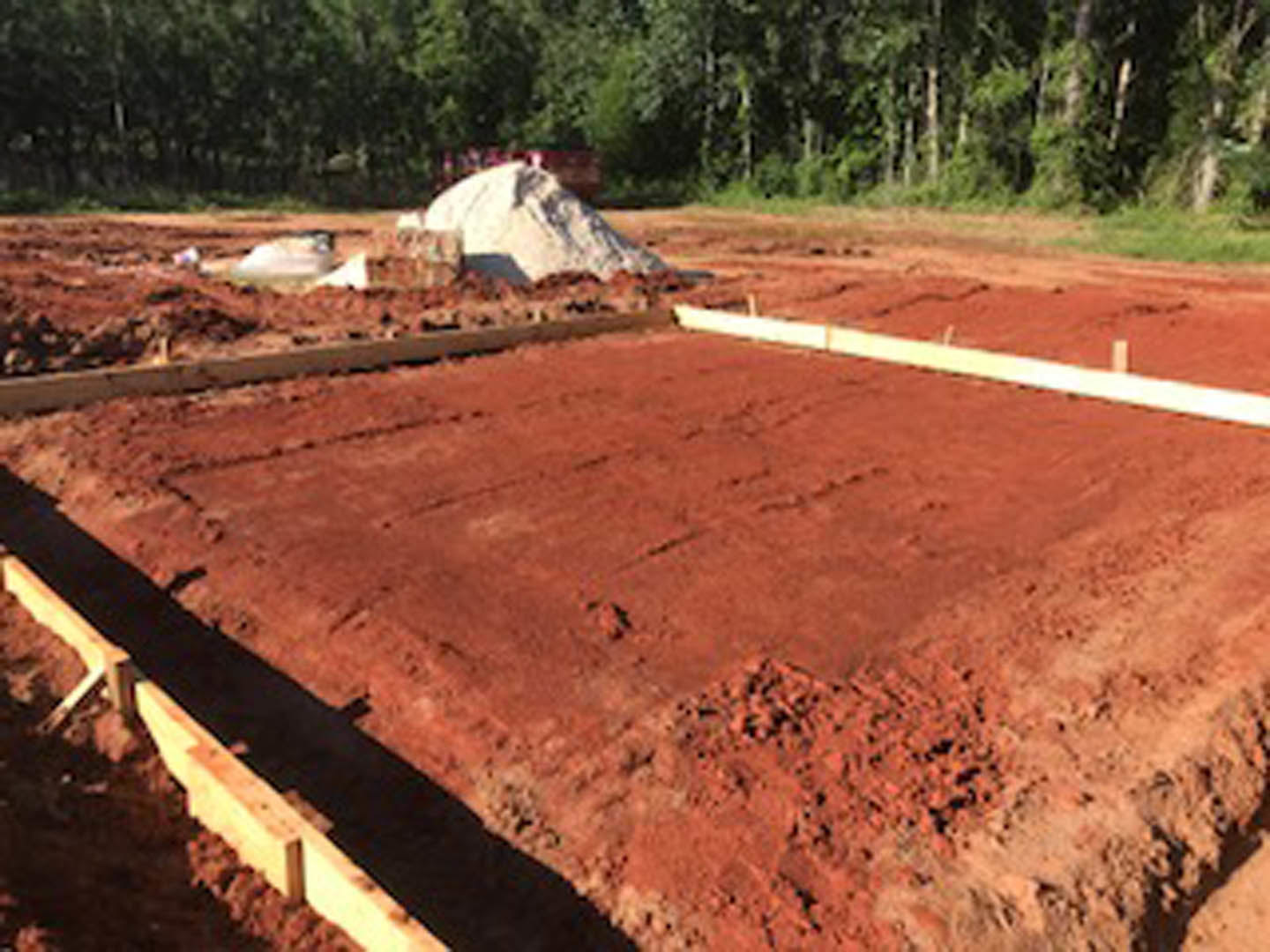 Concrete foundation under construction in red clay soil, wooden formwork outlining the perimeter, white chalk marking on ground, blurred tree and rock in background