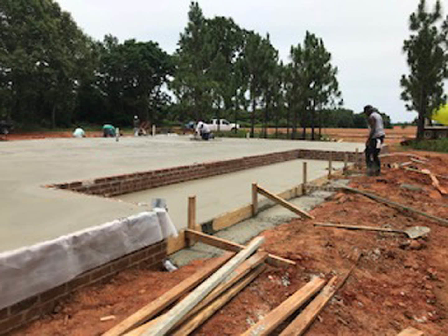 Partially built custom home foundation with exposed wood beams, concrete slab, and brick wall covered in white plastic; several workers on site, pile of lumber nearby, trees and