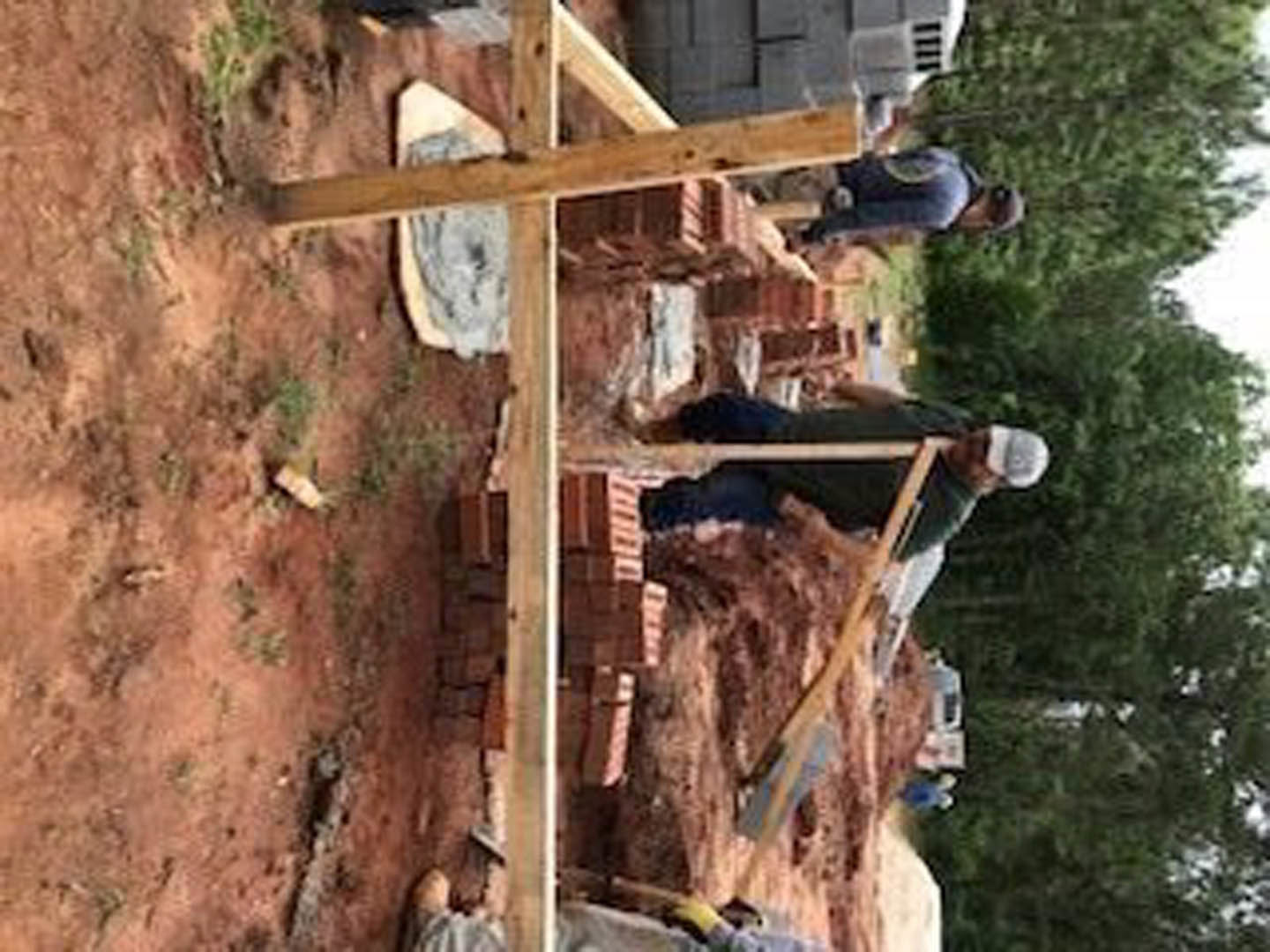 Framed wooden house under construction with several workers wearing casual clothing, surrounded by dirt ground and scattered building materials, trees in background