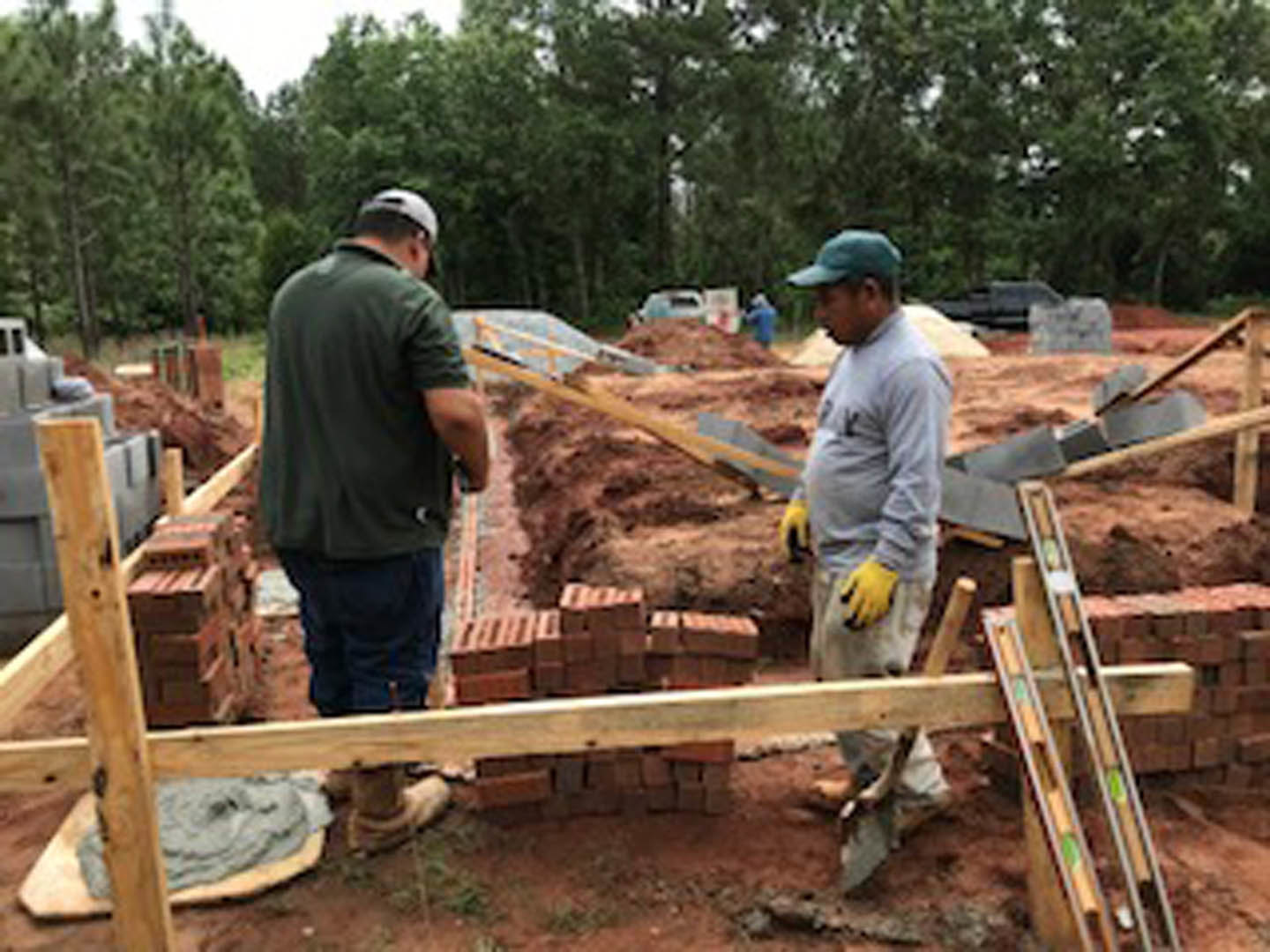 Several construction workers wearing gloves and work shirts assemble wooden posts and poles on a residential foundation, with soil and trees in the background.