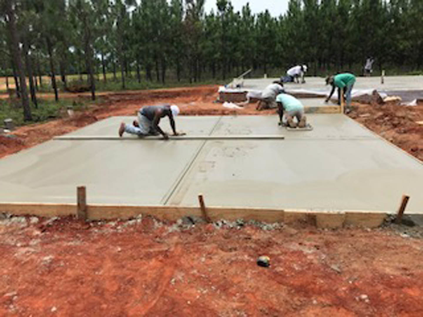 Workers pouring and smoothing concrete slab outdoors, surrounded by trees, wearing construction footwear.