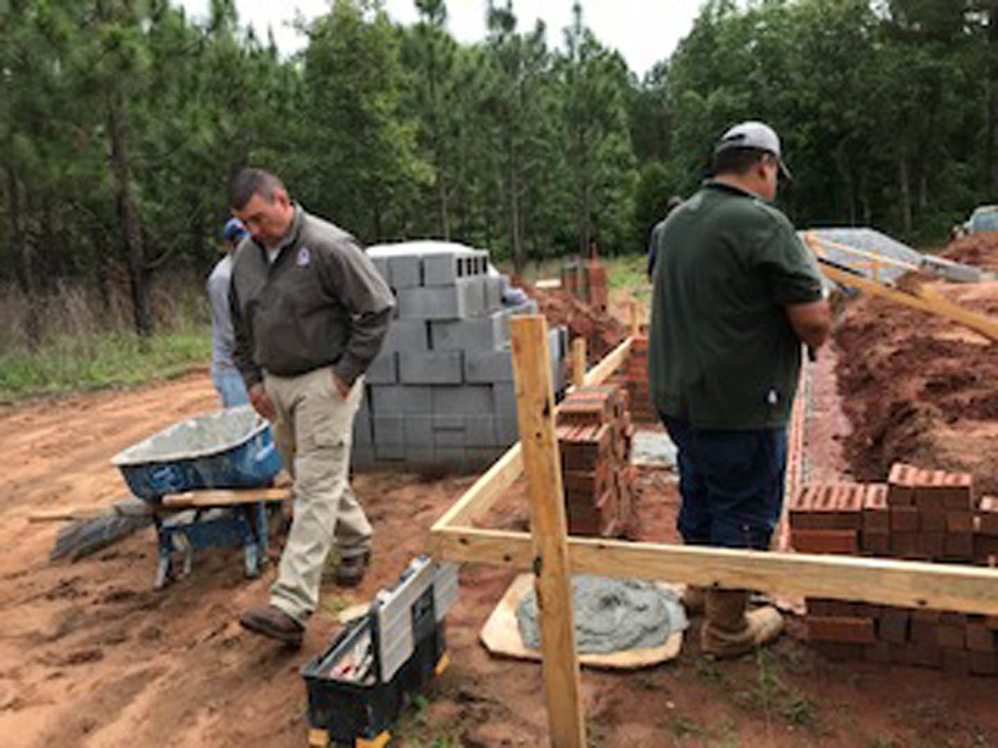 Framed house under construction with several men in work clothes standing on dirt foundation, open black plastic toolbox nearby, lumber and trees in background
