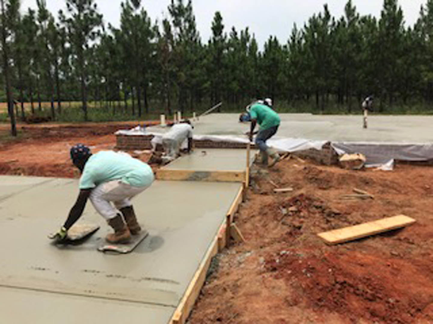 Crew pouring and leveling fresh concrete slab outdoors, surrounded by trees and construction materials, with workers wearing casual clothing and footwear.