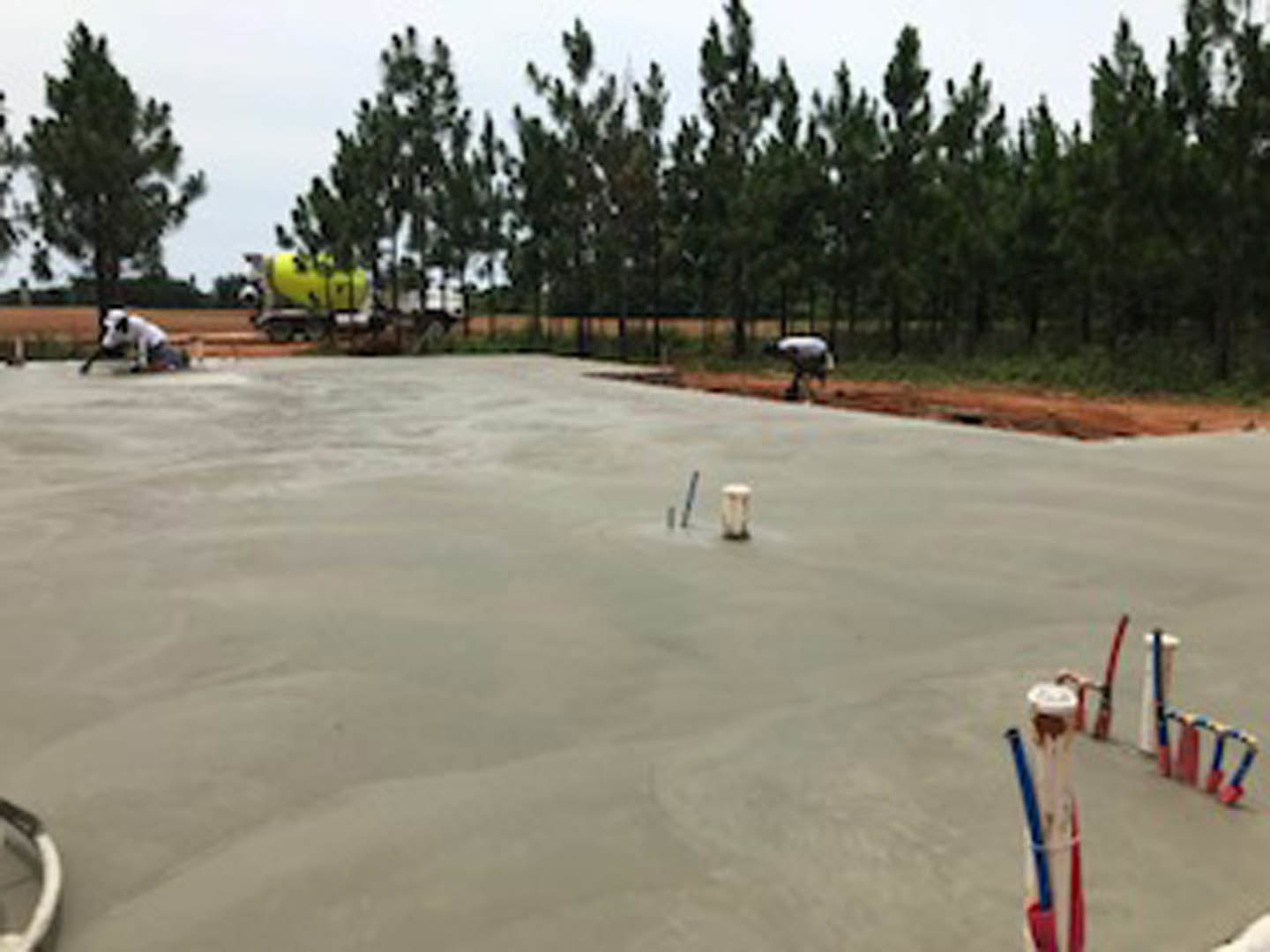 Freshly poured concrete slab with yellow cement mixer on roadside, surrounded by trees and open sky