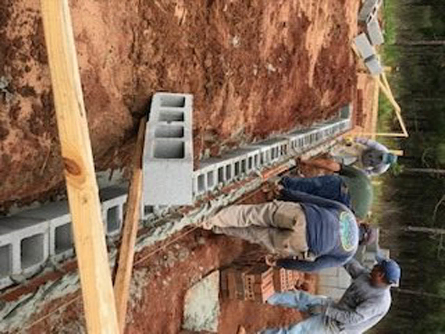 Men constructing a framed wall on a dirt lot, wooden posts and concrete blocks scattered, one person seated and another lying on the ground, casual work clothing, outdoor