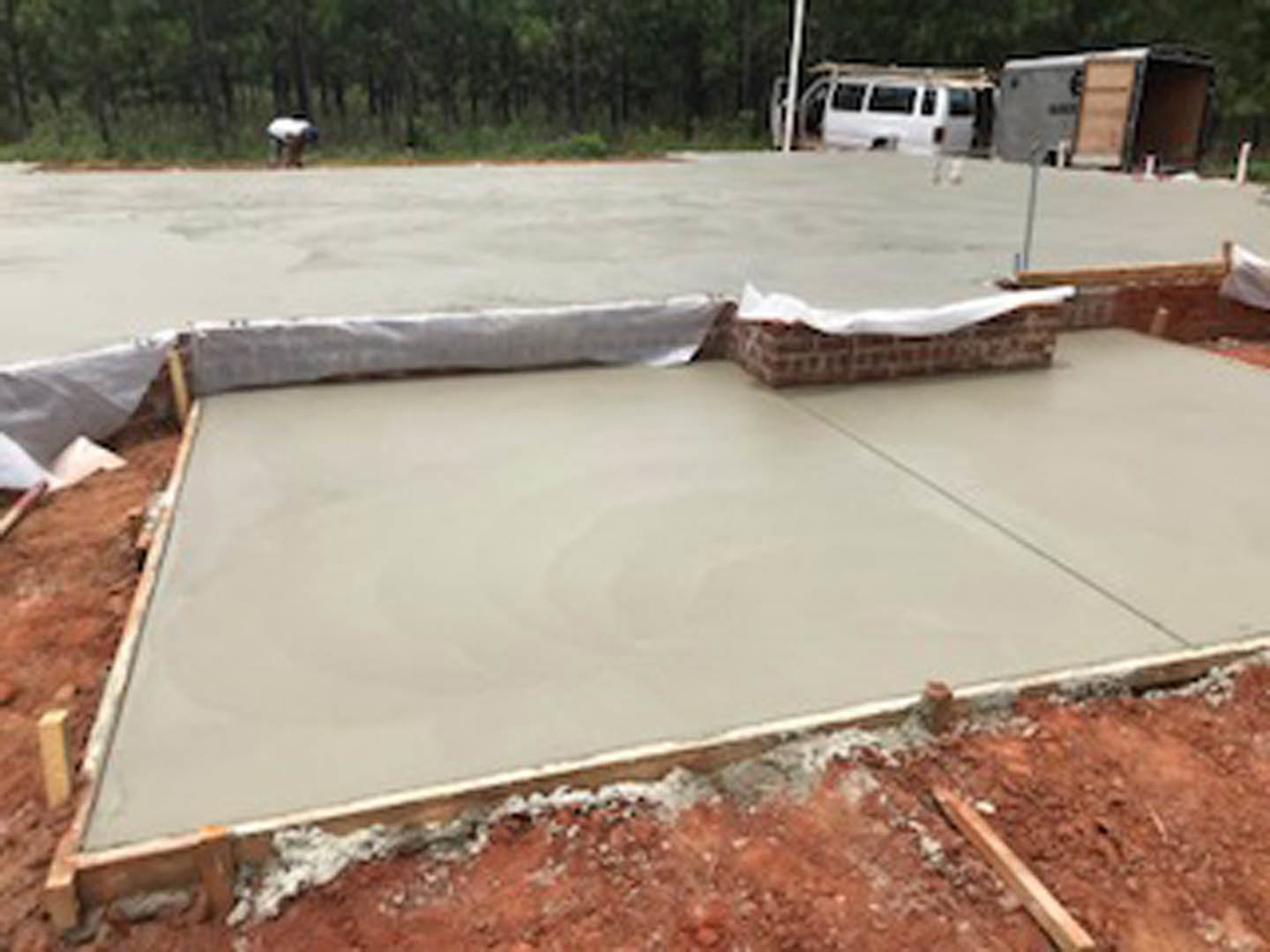 Freshly poured concrete slab surrounded by trees, with construction materials and a white van visible in the background