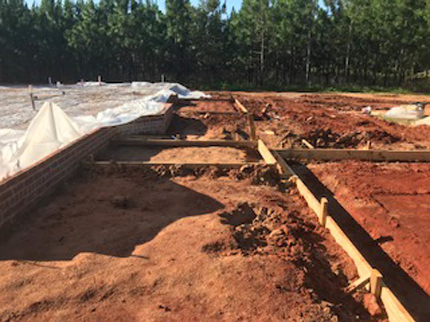 Brick wall under construction with exposed wooden beams, white tarp, and dirt foundation; trees and sky visible in the background.