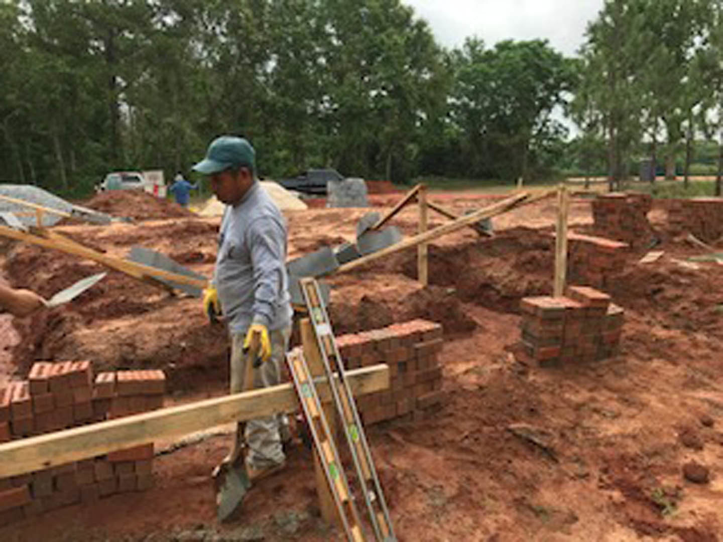 Construction worker in blue shirt, yellow gloves, and hat carrying wooden plank near soil and foundation with trees and parked cars in background