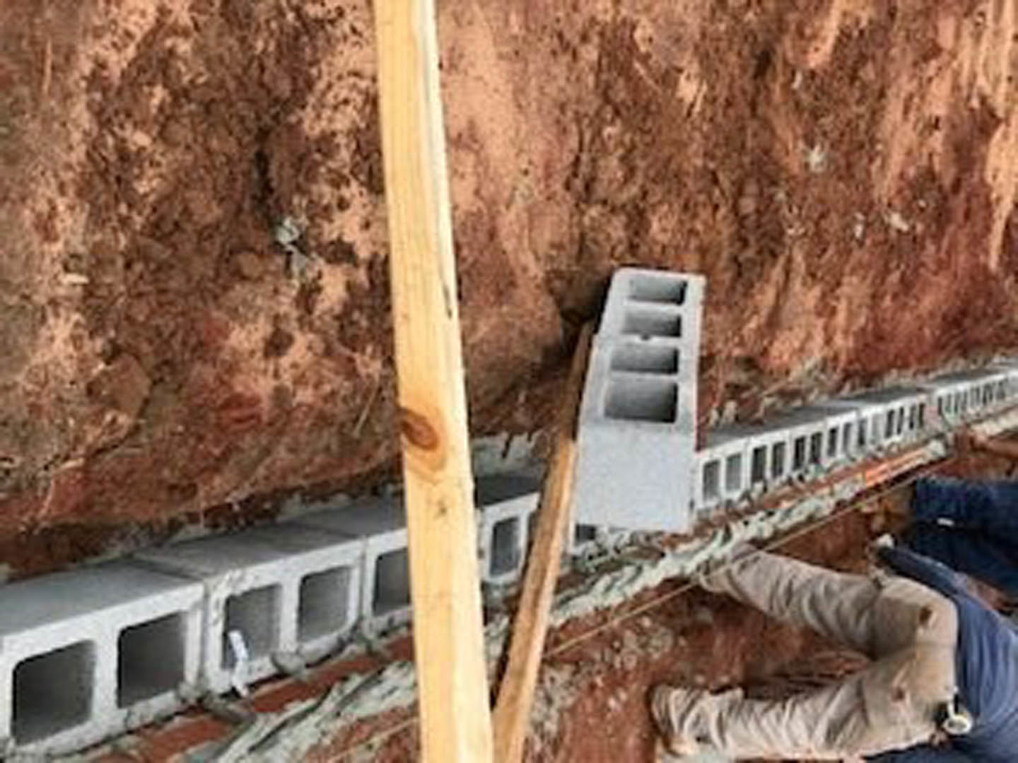 Worker in safety vest standing on dirt ground beside partially built concrete block wall and exposed wood framing of custom home exterior.