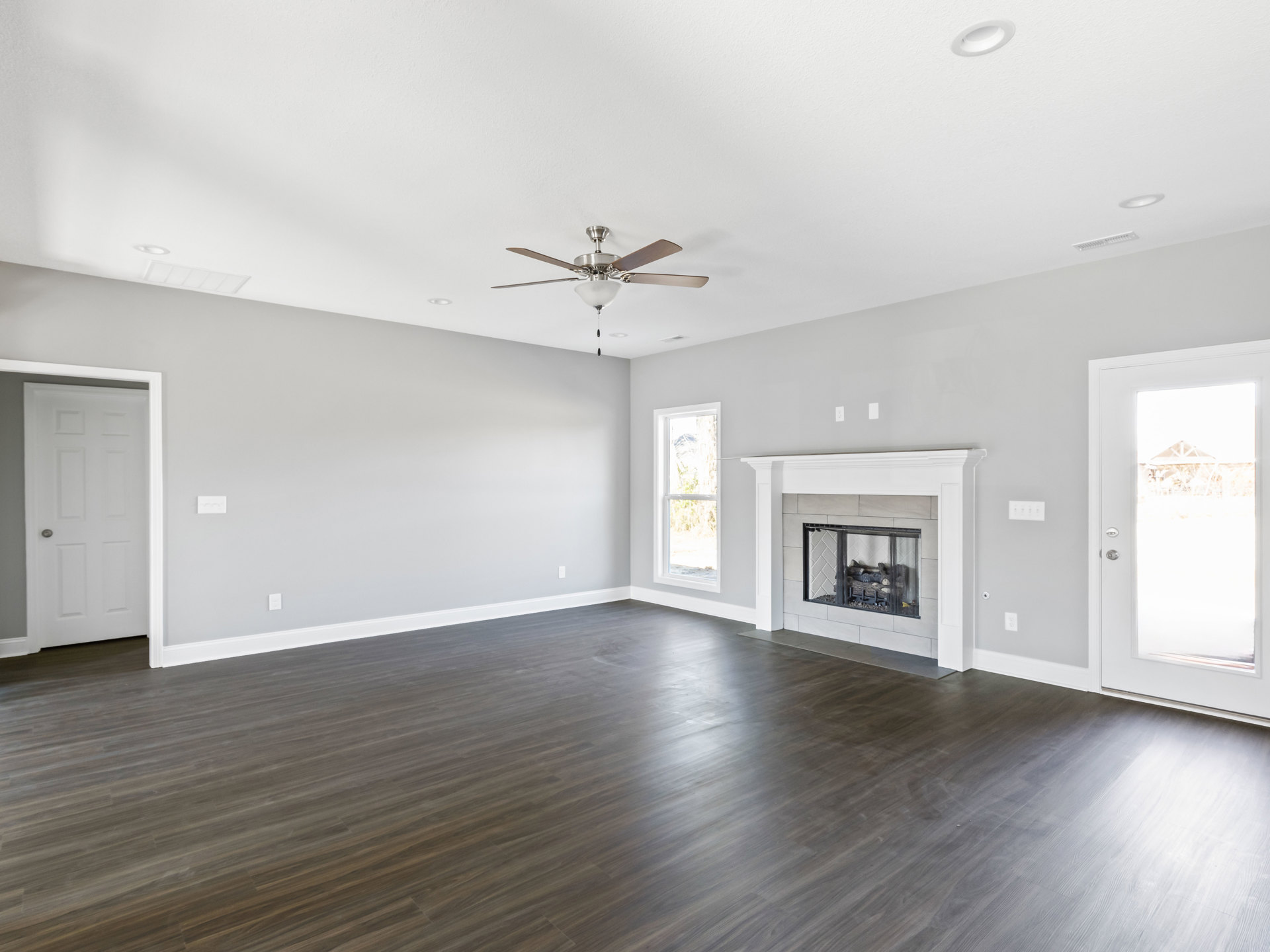 Living room with wood flooring, white plaster walls, glass-door fireplace, ceiling fan with light fixture, and glass door leading to outdoor gazebo