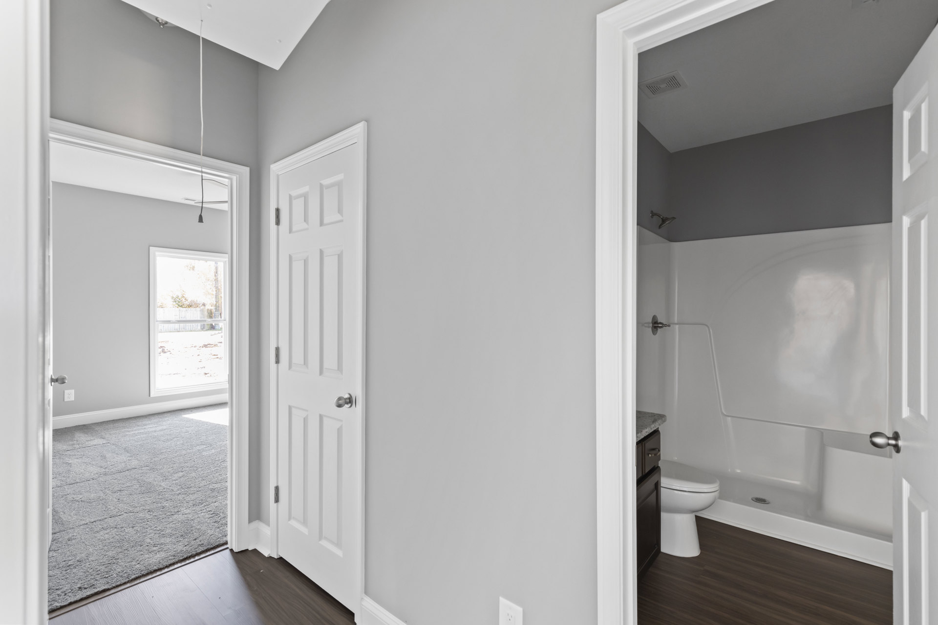 Bathroom with two white toilets, grey carpet flooring, white door with silver knob, ceiling vent, window overlooking fence and trees