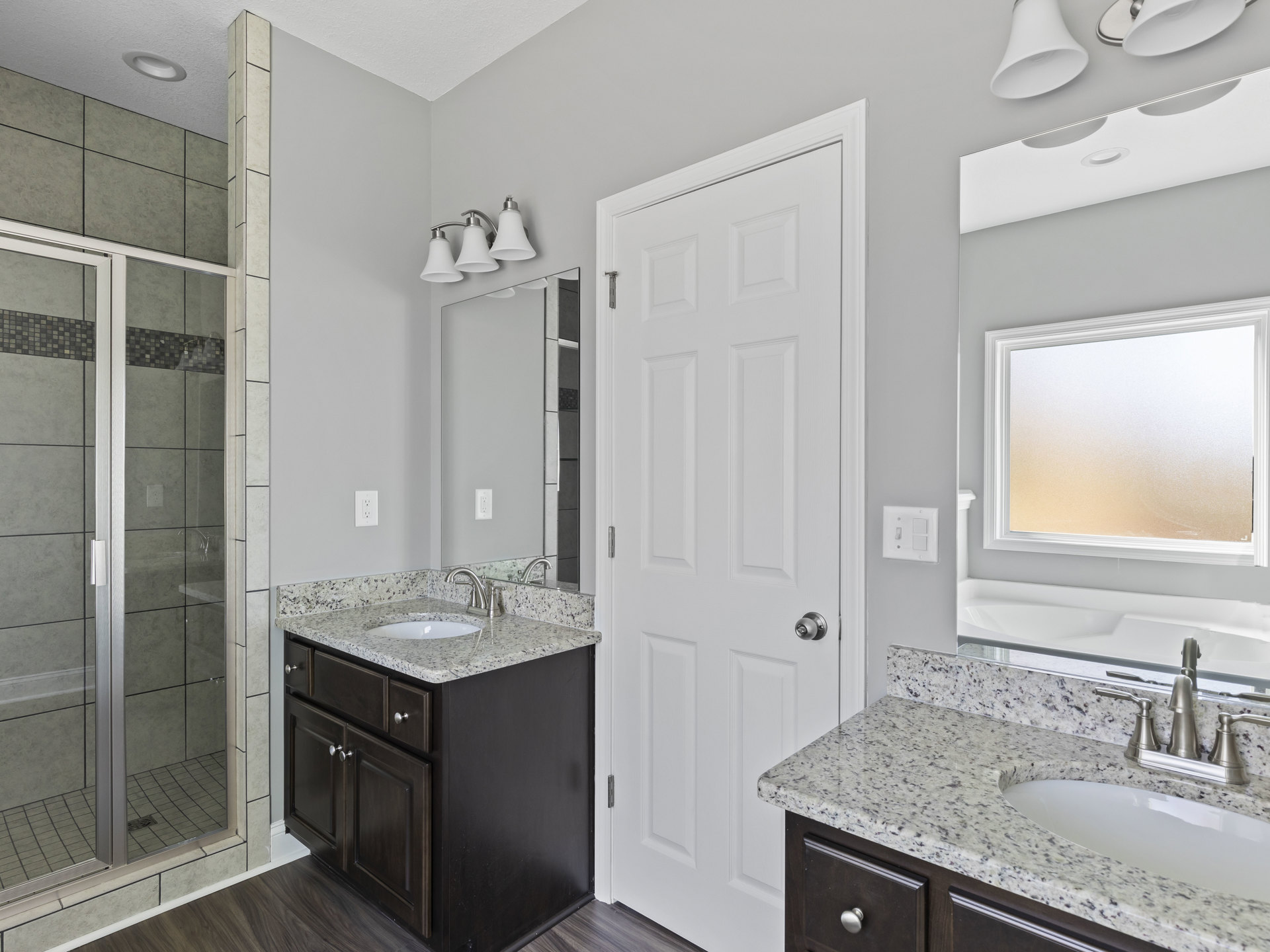 Bathroom with white paneled door, rectangular sink on stone countertop, glass shower enclosure, frosted window, white bell-shaped light fixture, and tiled walls.