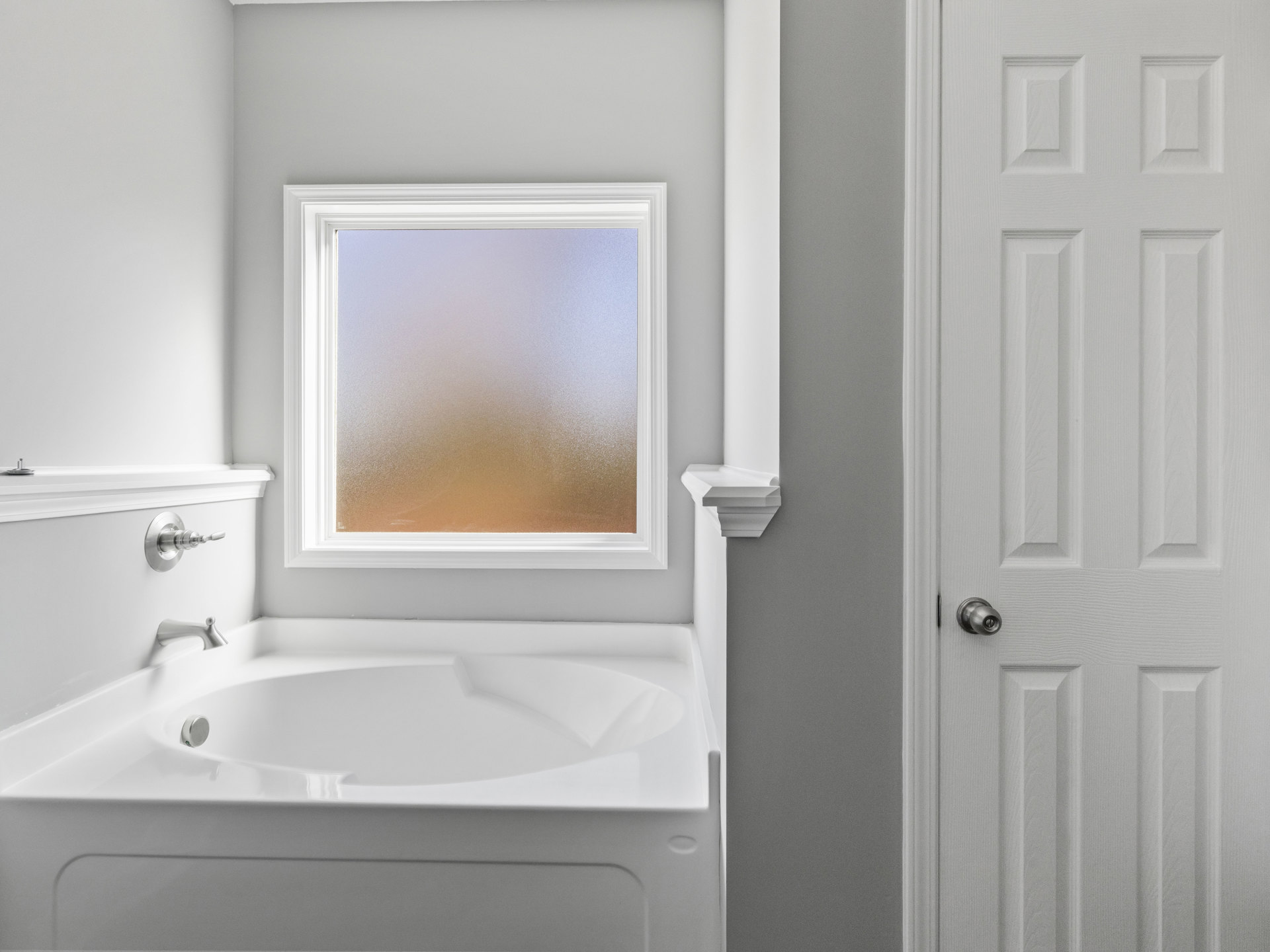 Freestanding white bathtub with chrome faucet beside frosted glass window, white tiled walls, and white door with silver knob
