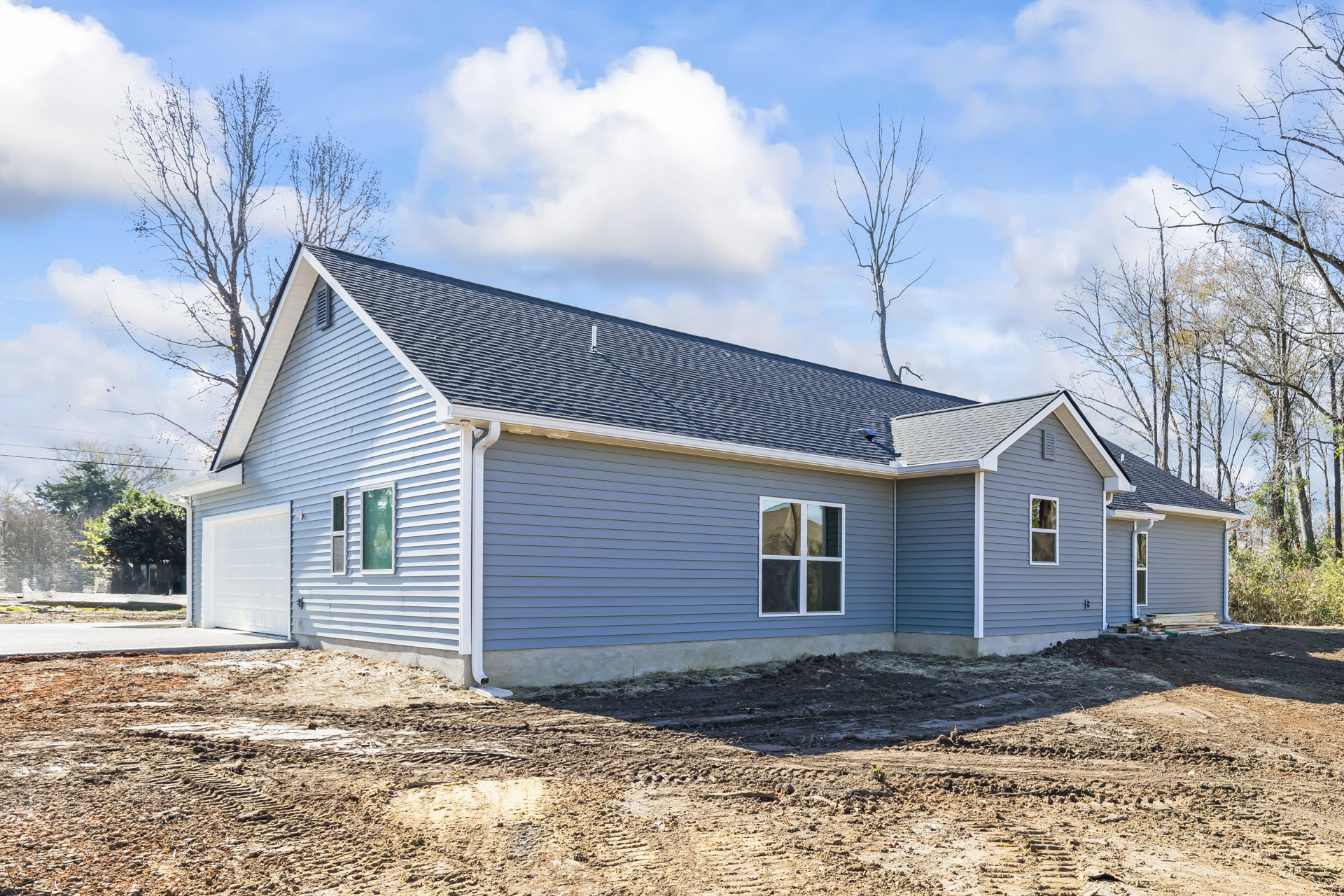 Framed custom home under construction with blue roof, large windows reflecting leafless tree, exposed dirt yard, scattered construction materials, and clear blue sky with clouds